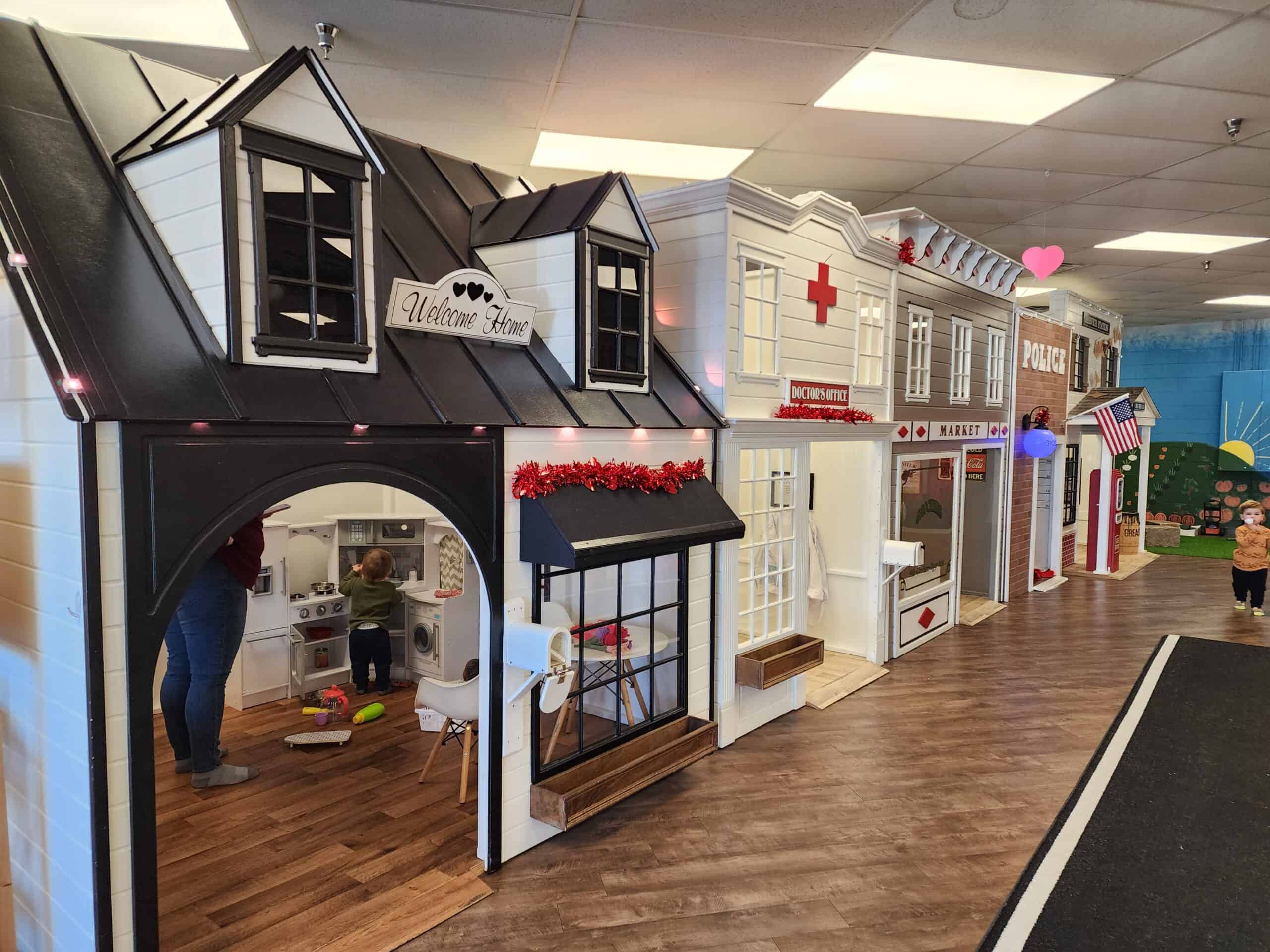 Interactive play area inside Piney Town Playhouse in Fuquay-Varina, NC, with a black playhouse featuring a 'Welcome Home' sign and a child playing in a realistic kitchen setup. Nearby are a doctor's office and a police station, all designed for educational play in a vibrant, small-scale town setting