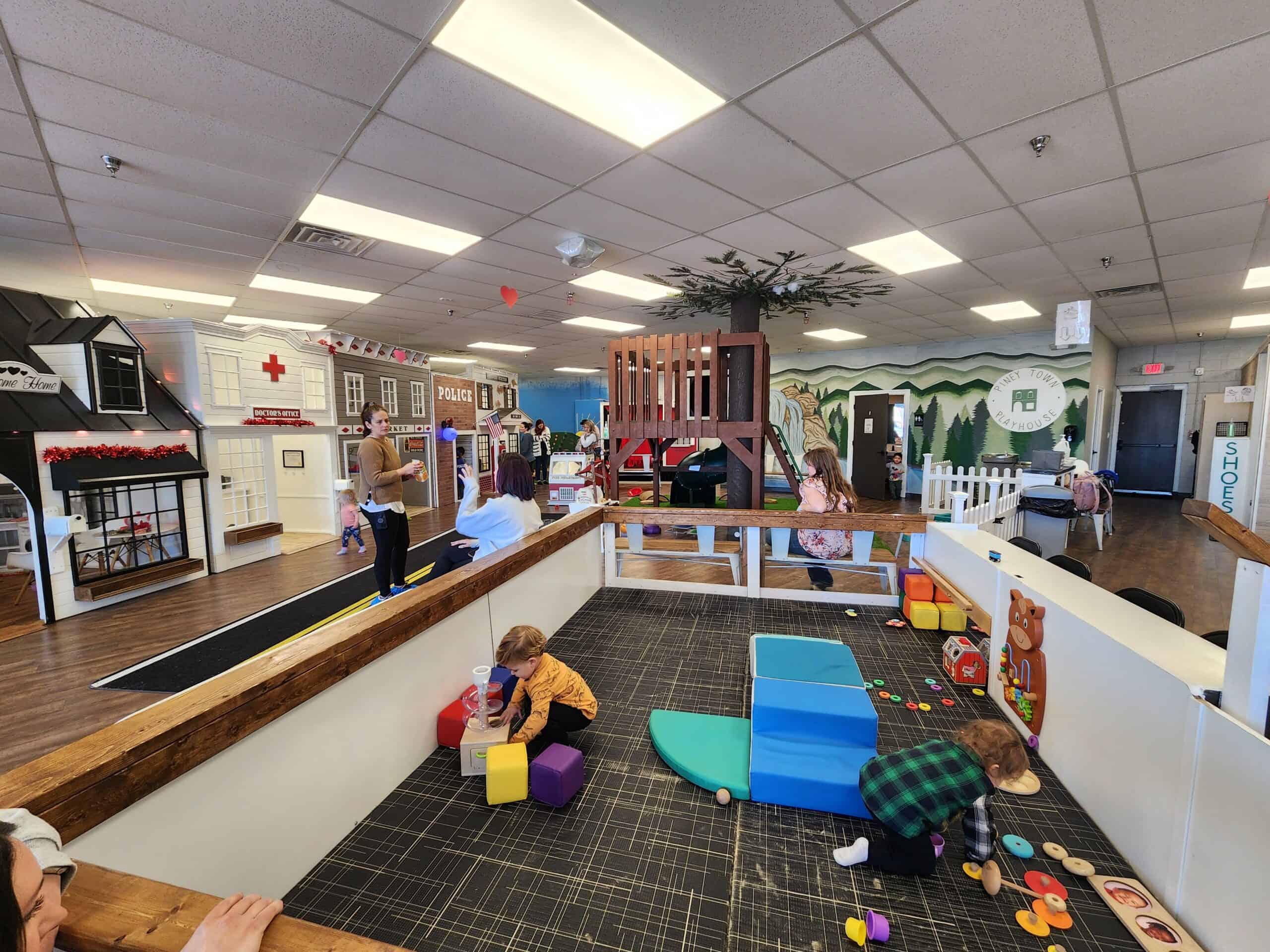 A bustling indoor play scene at Piney Town Playhouse in Fuquay-Varina, NC, with parents and children engaged in activities. The space features a miniature town with a black playhouse, a doctor's office, and a police station, fostering a fun and educational environment.