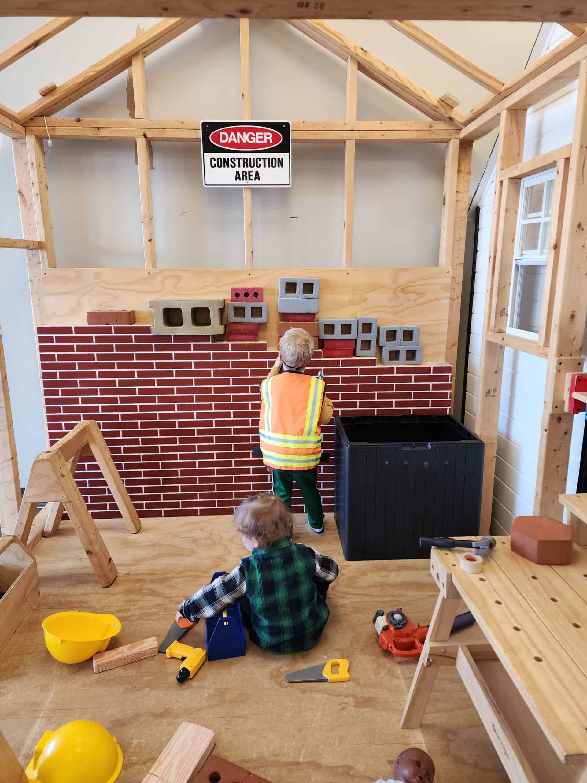 Young children in play construction vests engage in creative play at a 'Construction Area' within Piney Town Playhouse in Fuquay-Varina, NC, complete with toy tools, wooden blocks, and a brick wall backdrop, fostering a hands-on learning environment