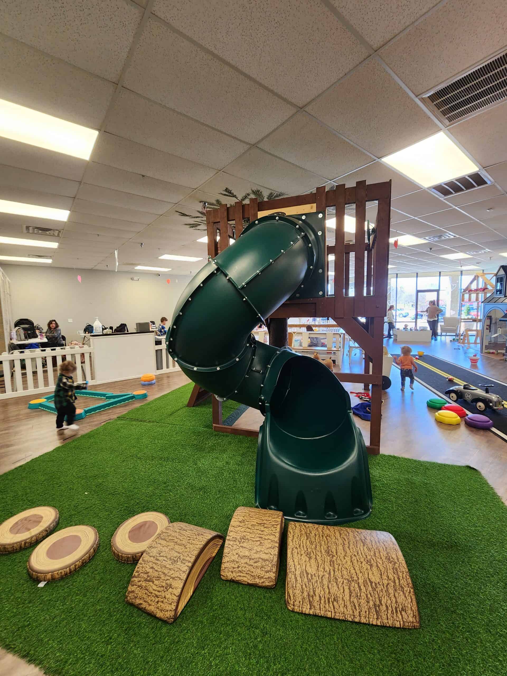 A vibrant green slide spirals down from a wooden play structure at Piney Town Playhouse in Fuquay-Varina, NC, with a set of log-like stepping stones leading up to it on the artificial grass. Children enjoy playtime in the background with a view of the indoor play town