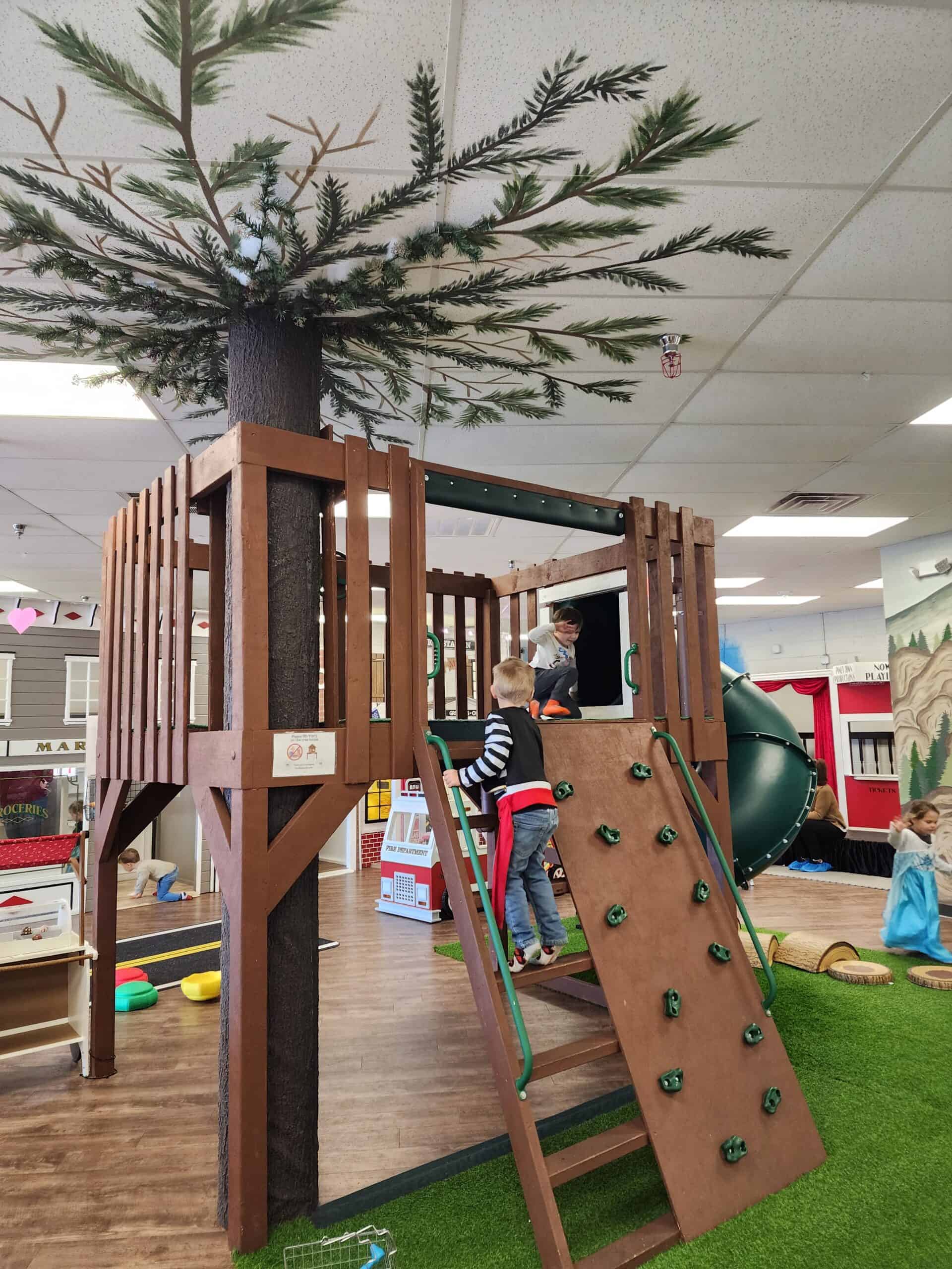 Children play on a wooden climbing structure with a rock wall and a slide, designed to resemble a treehouse, at Piney Town Playhouse in Fuquay-Varina, NC. The indoor play area also features artificial turf and a backdrop of colorful, kid-sized storefronts.