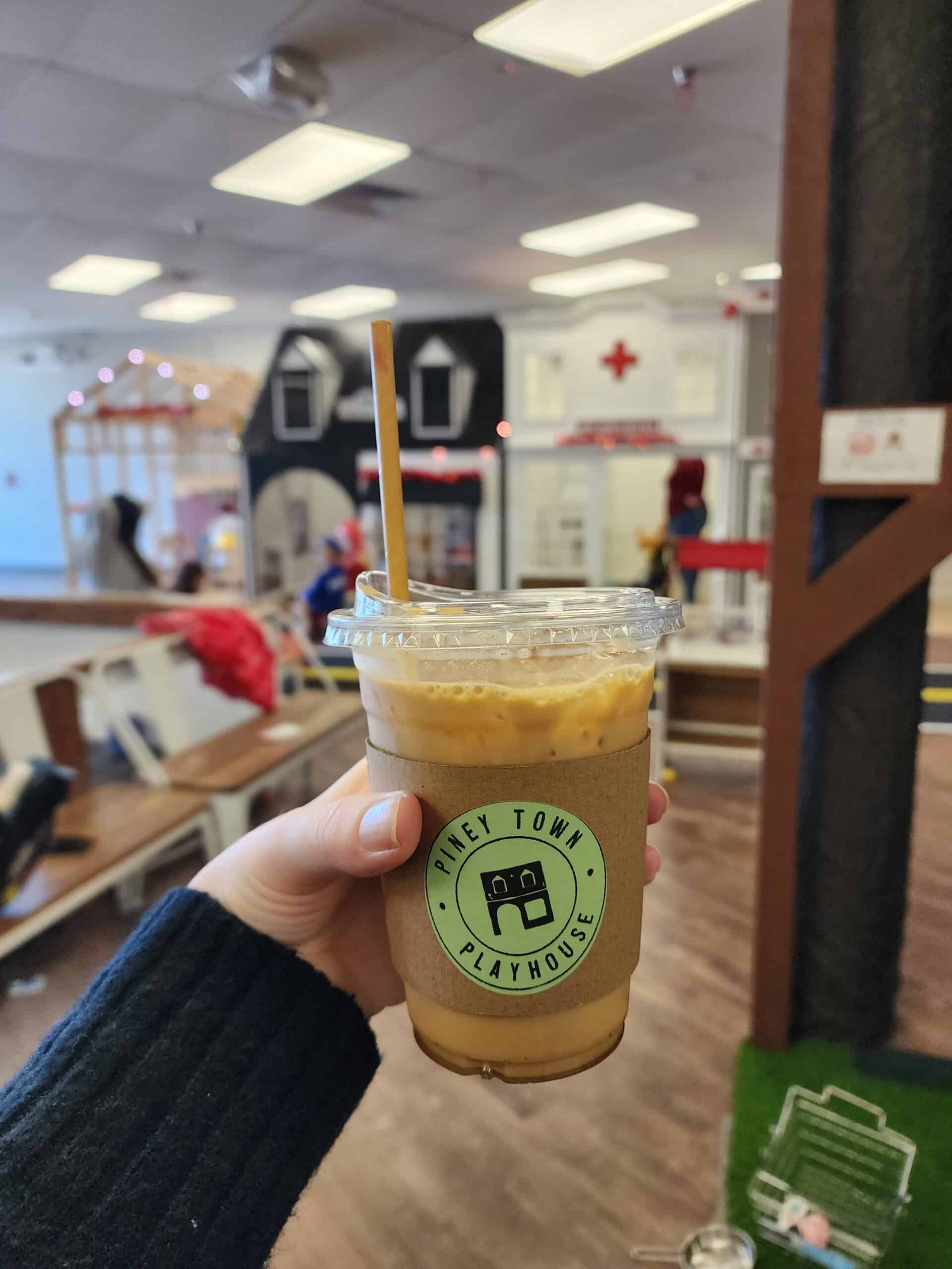 Close-up of a hand holding a Piney Town Playhouse branded iced coffee cup with a paper straw, in focus against a blurred background of the playful indoor town setting in Fuquay-Varina, NC