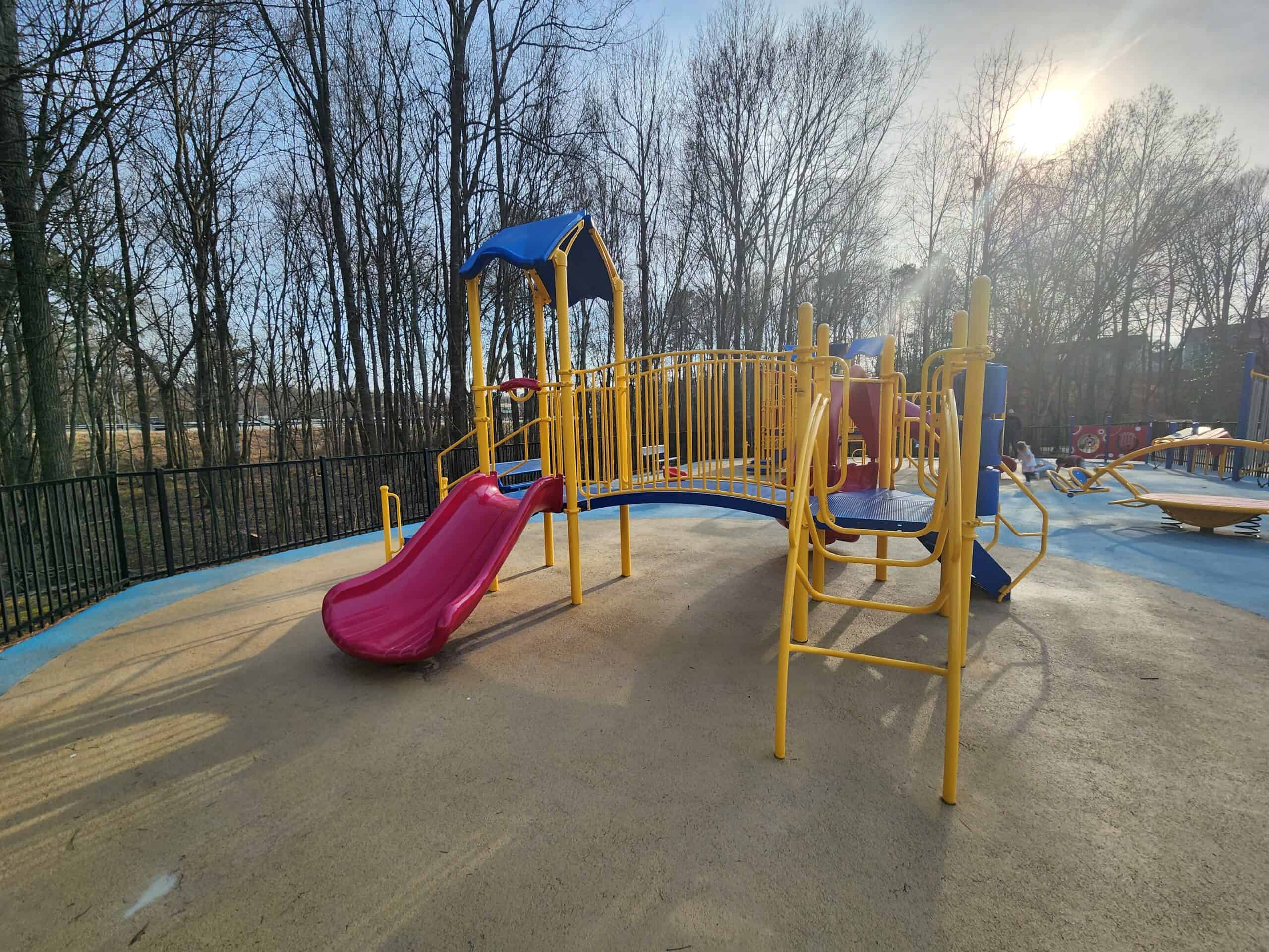 A fenced playground in Morrisville, NC, featuring bright yellow and blue play structures with red slides and bridges, set on a soft, colorful surface. The playground is surrounded by leafless trees on a clear, sunny day.