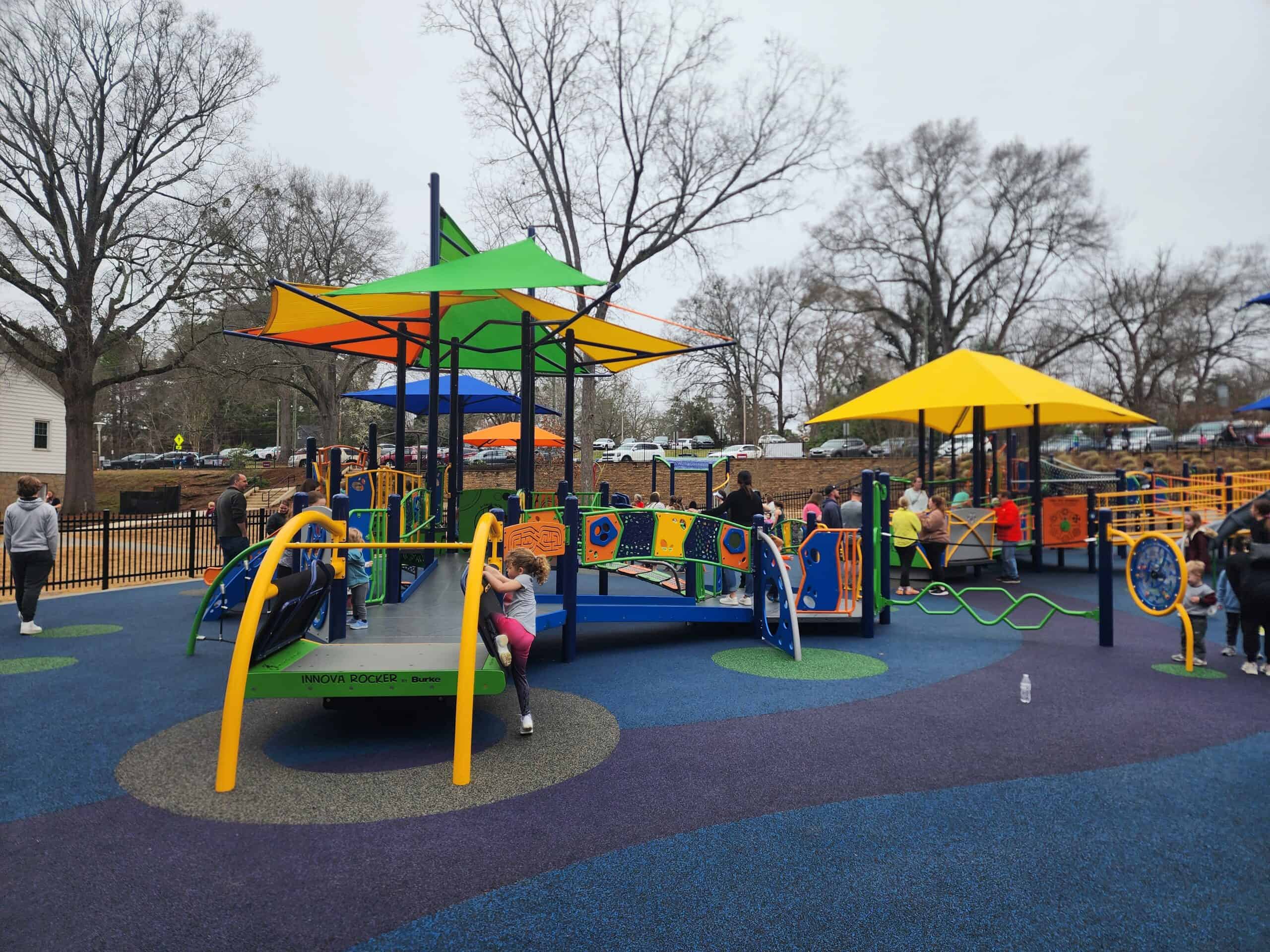 A fenced playground in Wake Forest, NC, featuring vibrant play structures with colorful shade canopies, including climbing elements and interactive panels. Children are playing on the soft, blue and green rubberized surface under the watchful eyes of parents on a cloudy day.