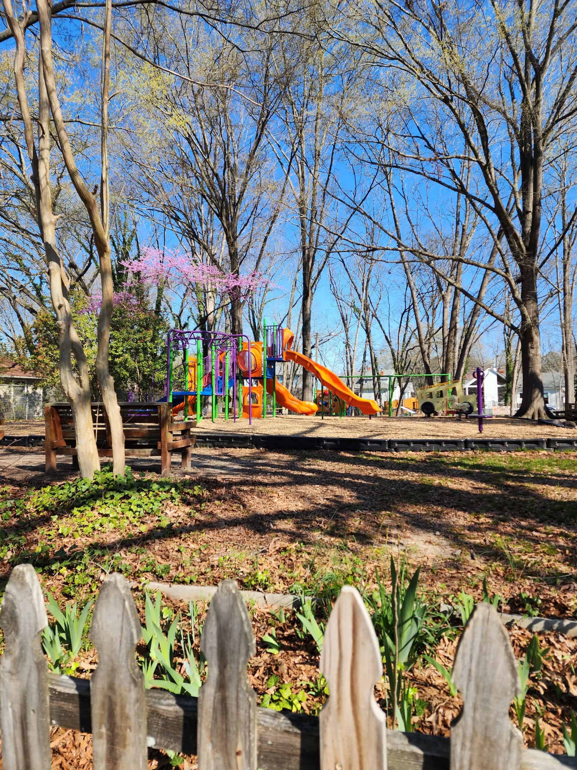 A fenced playground in Durham, NC, featuring colorful play structures with orange slides, surrounded by tall trees and blooming flowers. The playground is set on a woodchip surface with benches and a picket fence in the foreground on a clear, sunny day