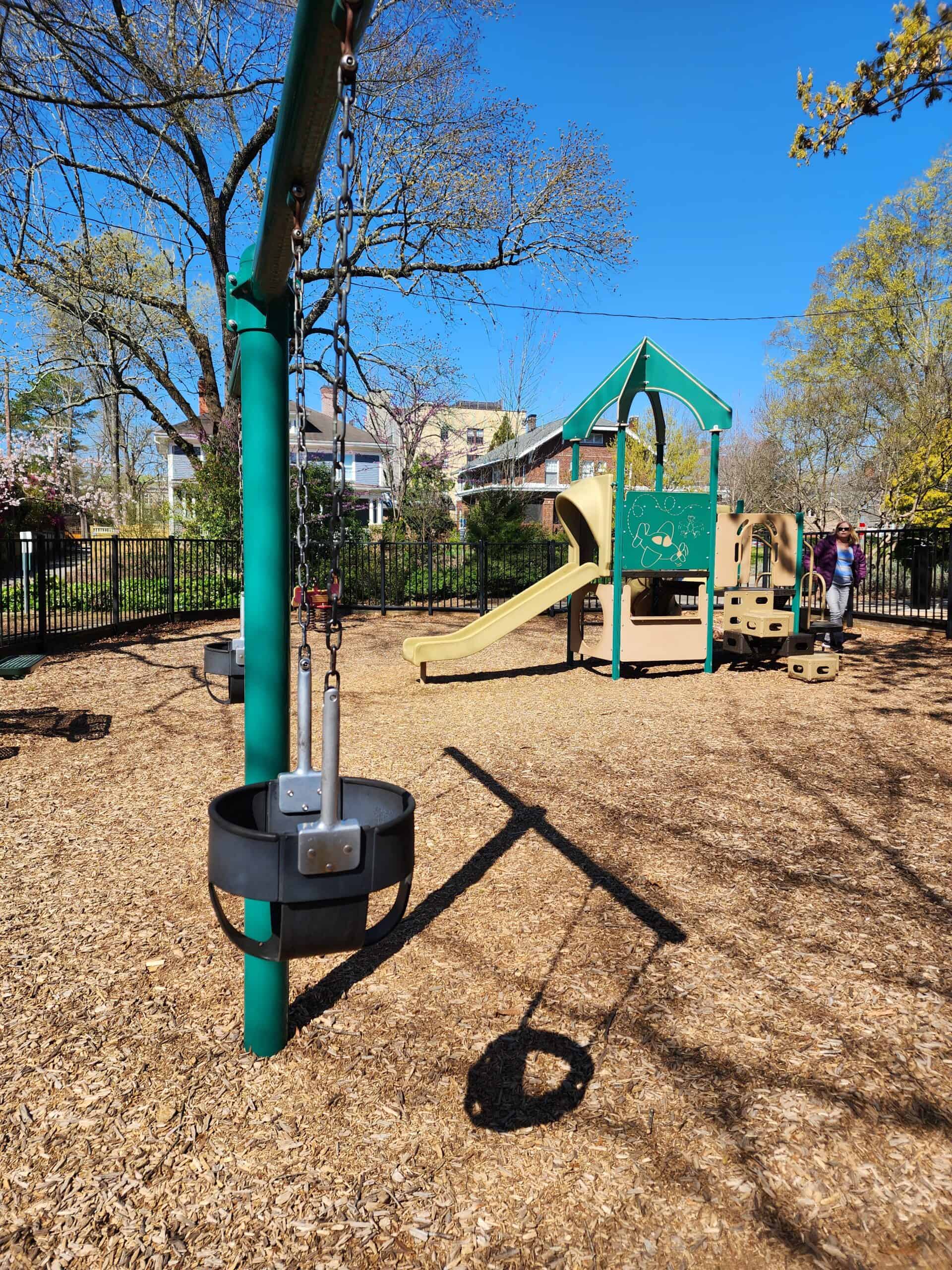 A fenced playground featuring a beige and green play structure with slides, climbing elements, and baby swings, set on a woodchip surface. The playground is surrounded by trees and residential buildings on a clear, sunny day