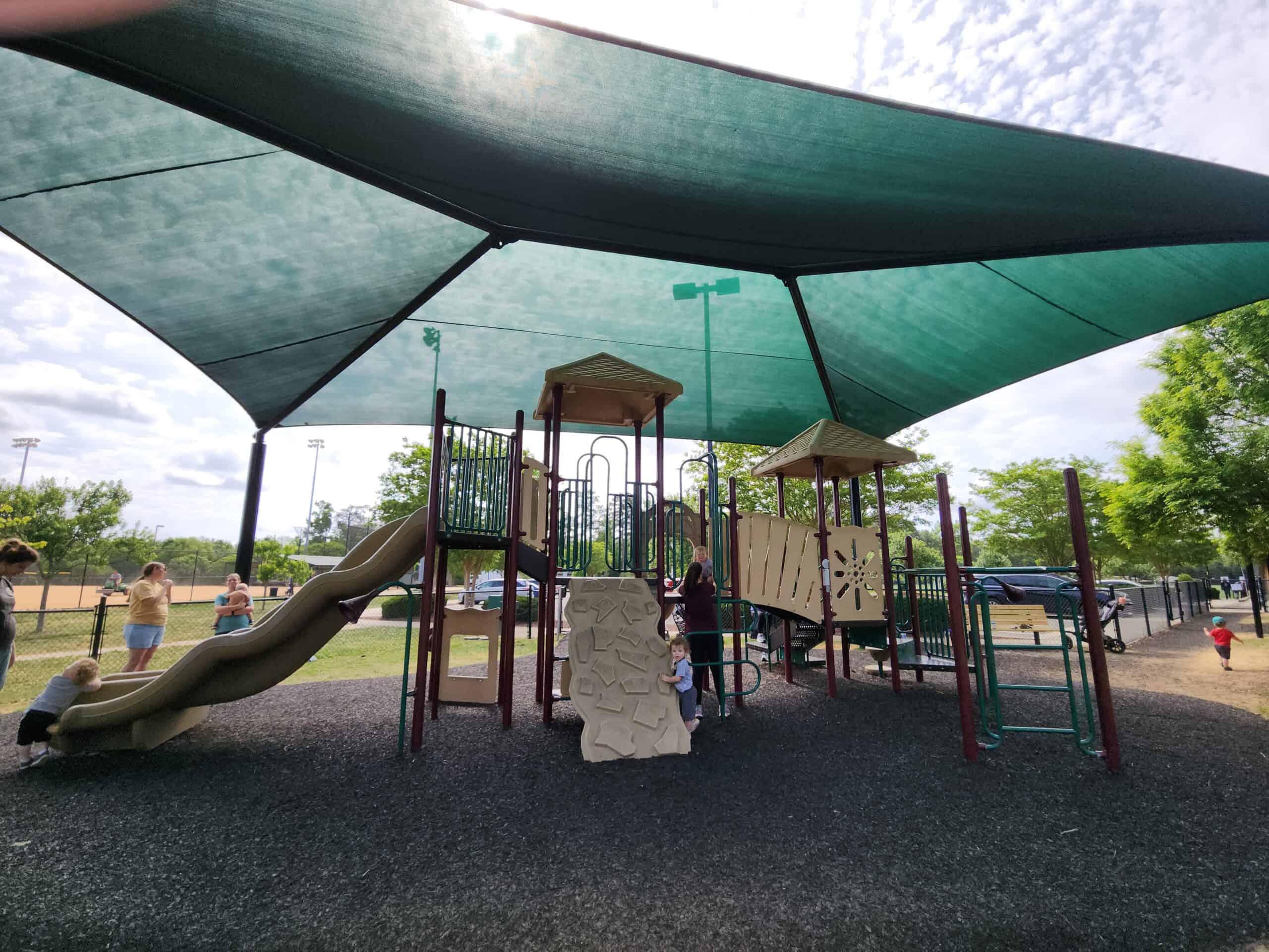 A fenced playground in Apex, NC, featuring shaded play structures with beige and brown slides, climbing elements, and bridges, set on a rubberized surface. Parents and children are enjoying the play area under a large green canopy on a partly cloudy day.