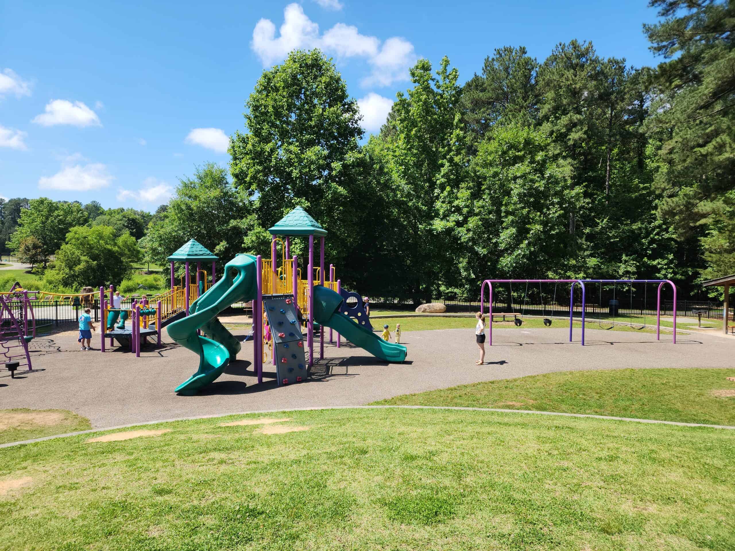 A fenced playground in Chapel Hill, NC, featuring vibrant green and purple play structures, including slides, climbing walls, and swings. Children are playing under a bright blue sky with lush green trees in the background