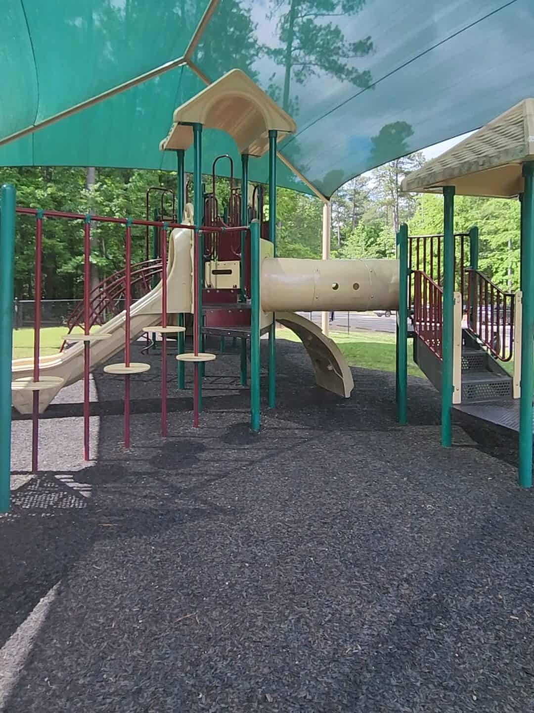 A shaded fenced playground featuring beige and maroon play structures, including slides, climbing elements, and platforms, set on a soft, rubberized surface. The playground is surrounded by green trees, providing a safe and comfortable play area for children