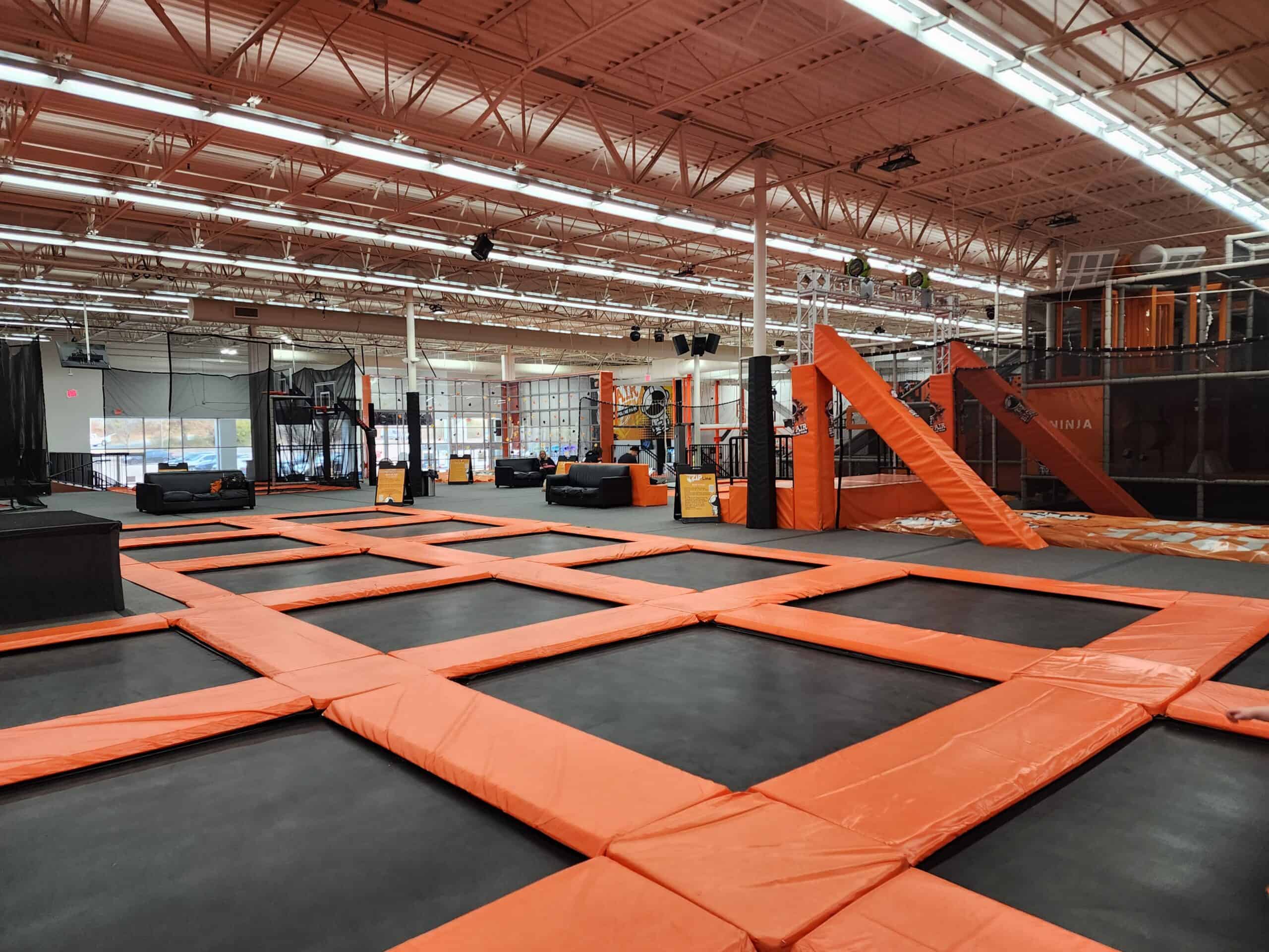 A spacious indoor trampoline park in Raleigh, NC, featuring a large grid of interconnected trampolines with black jumping surfaces and orange padding. The park includes various attractions like a basketball area, ninja course, and cushioned seating areas, providing a fun and dynamic environment for visitors.