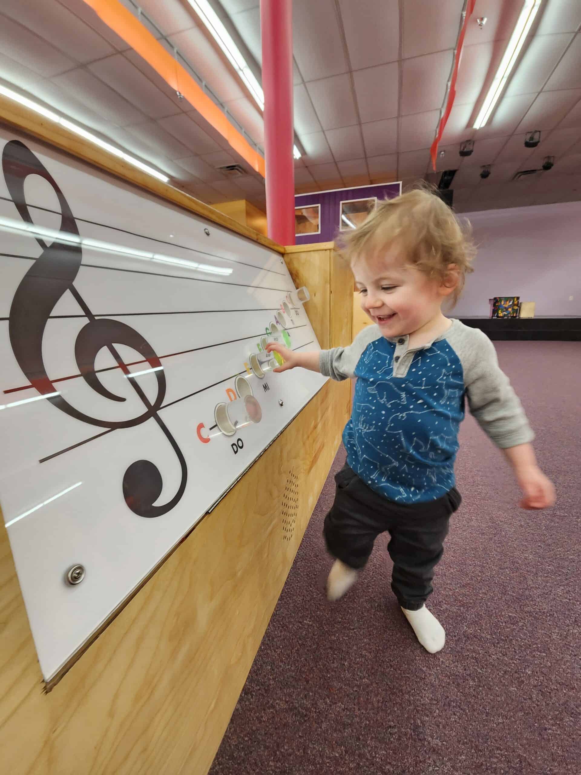 A smiling toddler interacts with a musical wall display featuring a large treble clef and a series of buttons labeled with musical notes such as "C" and "D." The child appears engaged and happy in the colorful, indoor play area.