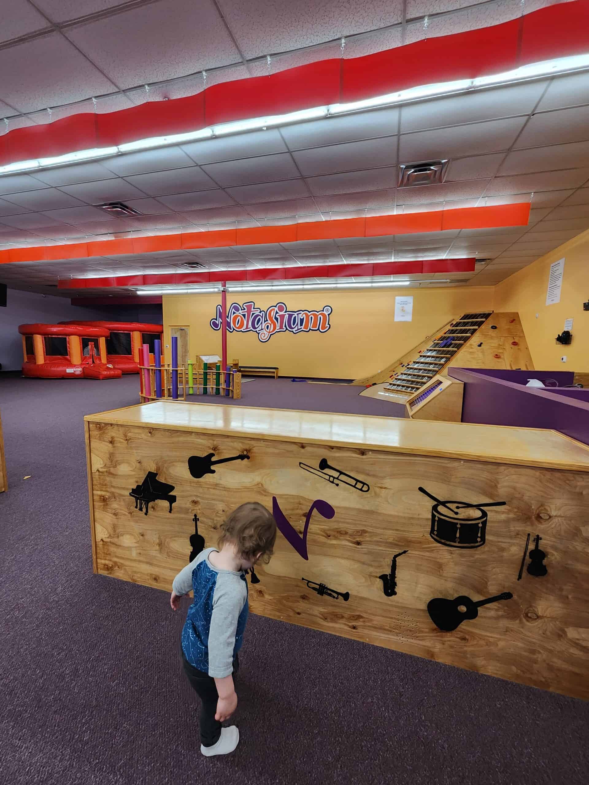
A small child stands in an indoor play area with the sign "Notasium" in the background. The space features colorful play equipment and a wooden partition decorated with silhouettes of musical instruments.