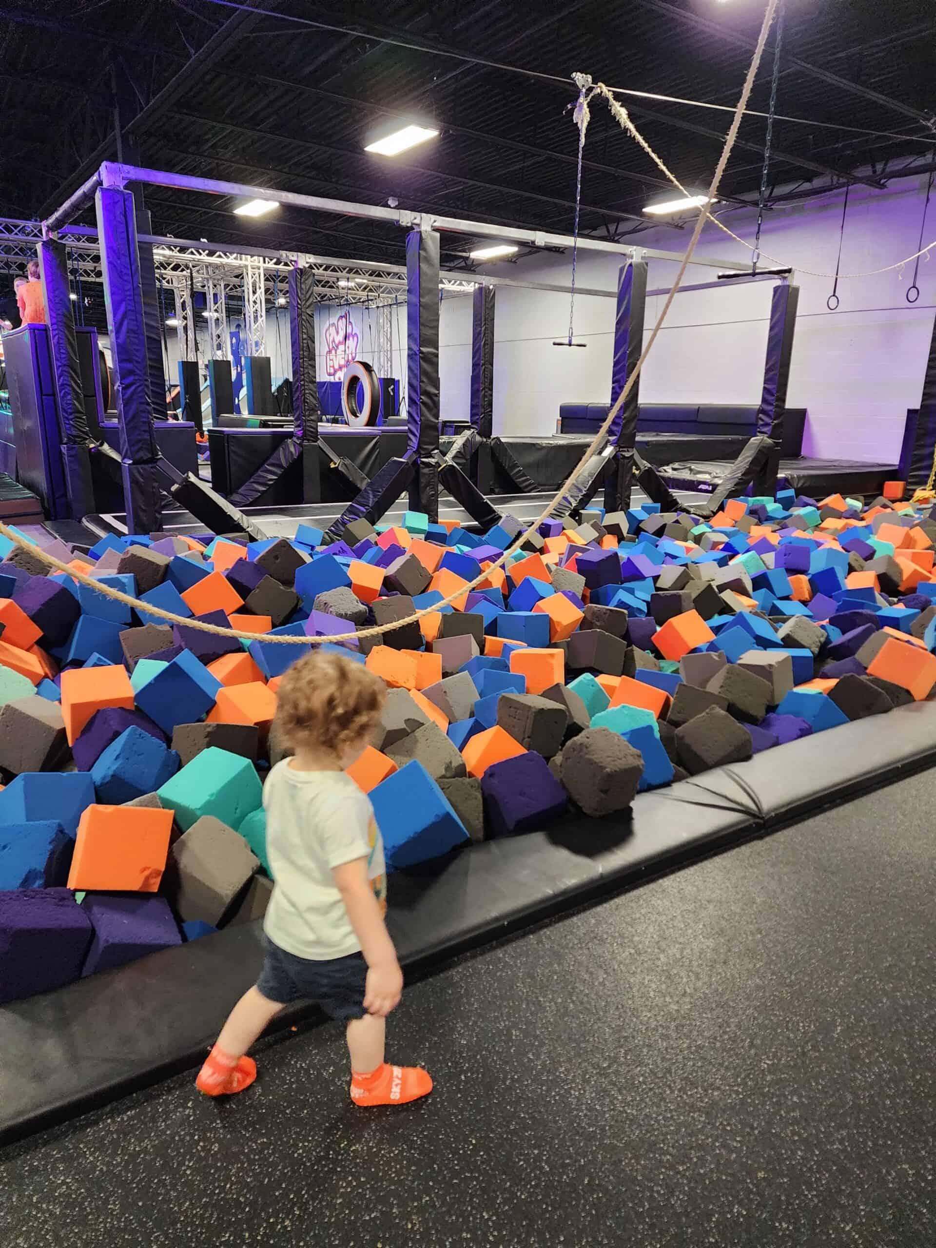 A young child walks past a foam pit filled with colorful foam cubes in an indoor adventure park. The park features ropes, climbing structures, and various obstacles, providing a vibrant and engaging space for active play.