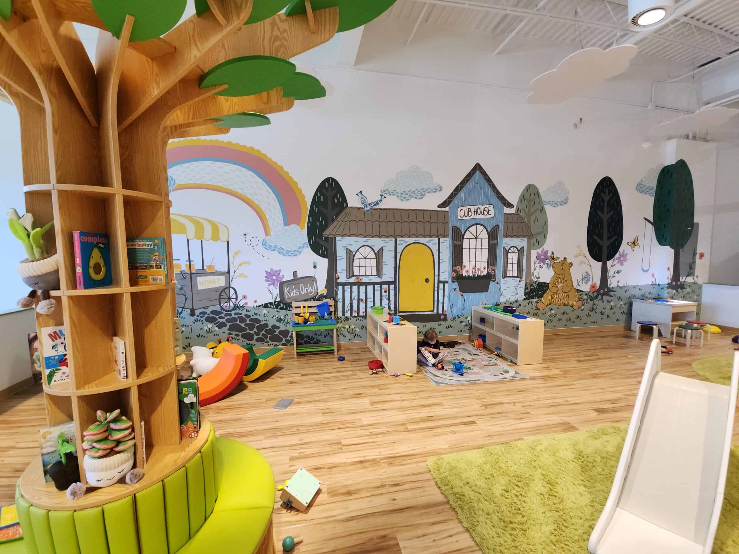 A colorful indoor playground features a whimsical mural of a clubhouse and a honey cart, with a tree bookshelf stocked with children's books. There is a play area with toys scattered on the floor, a small slide, and bright green rugs, creating a playful and inviting atmosphere for kids in Clayton, NC.
