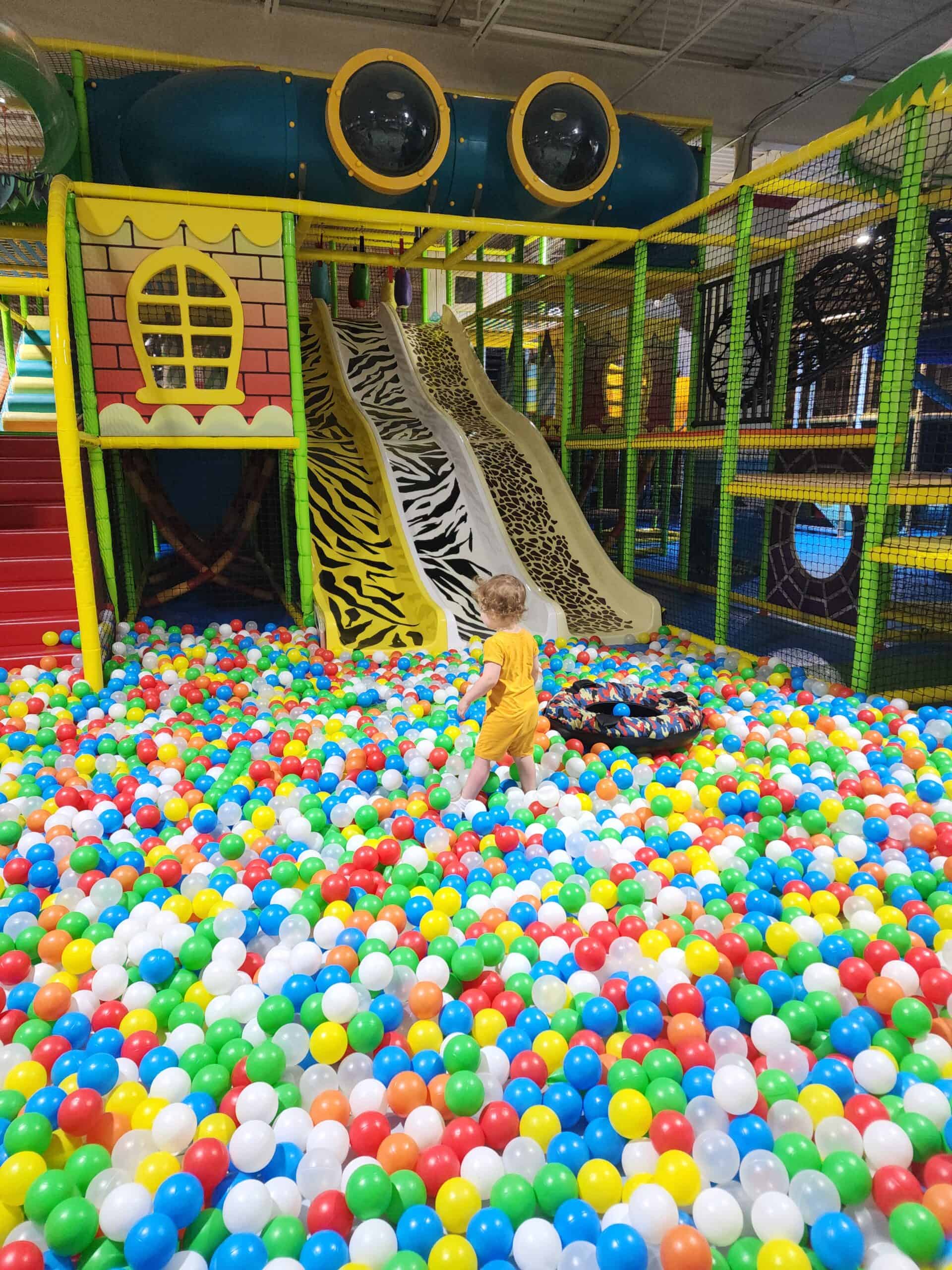 a huge colorful ballpit surrounds a soft play area for kids with animal print slides heading into it. A toddler walks through the ball pit toward the play area