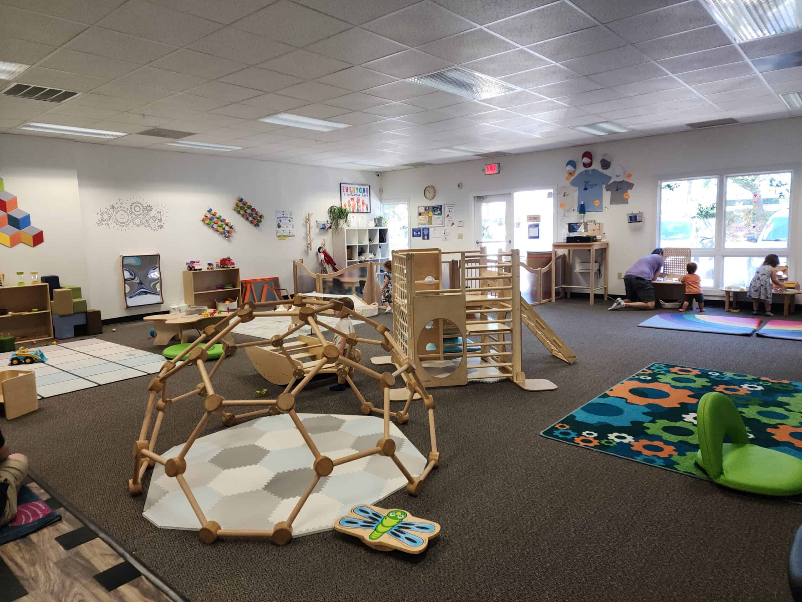 A spacious indoor play area in Durham is filled with various wooden play structures, toys, and colorful mats. Children and an adult engage in different activities, with natural light streaming through large windows. The room has a vibrant, welcoming atmosphere, featuring educational wall decorations and organized play zones.