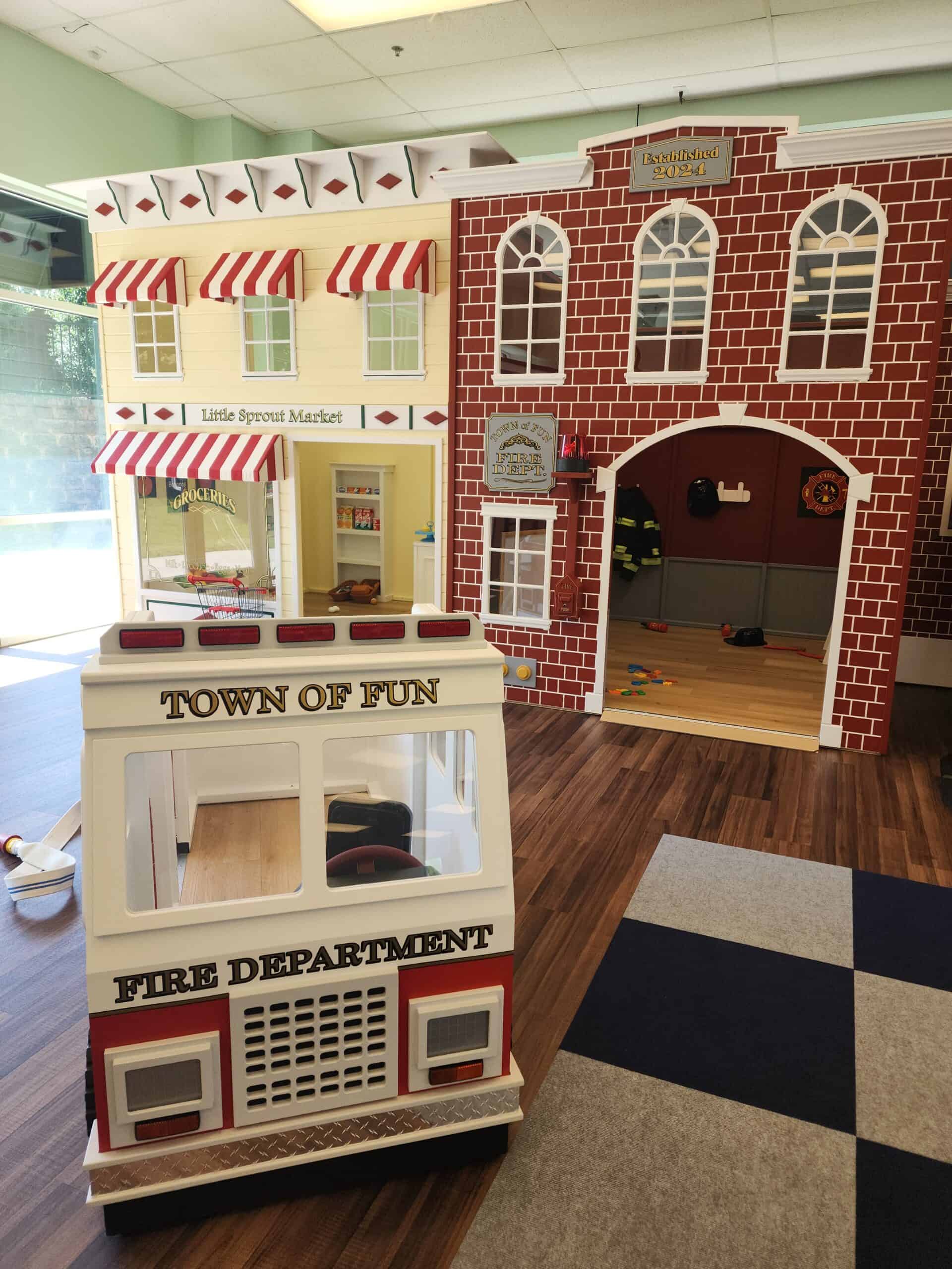 This image captures a playful, miniature town setup for children, featuring a "Town of Fun" fire department truck prop and themed storefronts. On the left is the "Little Sprout Market," styled with red-and-white striped awnings, and on the right is a brick-style fire department building labeled "Established 2024." The wooden floors and colorful props create an immersive, role-play environment designed to spark imagination and pretend play.