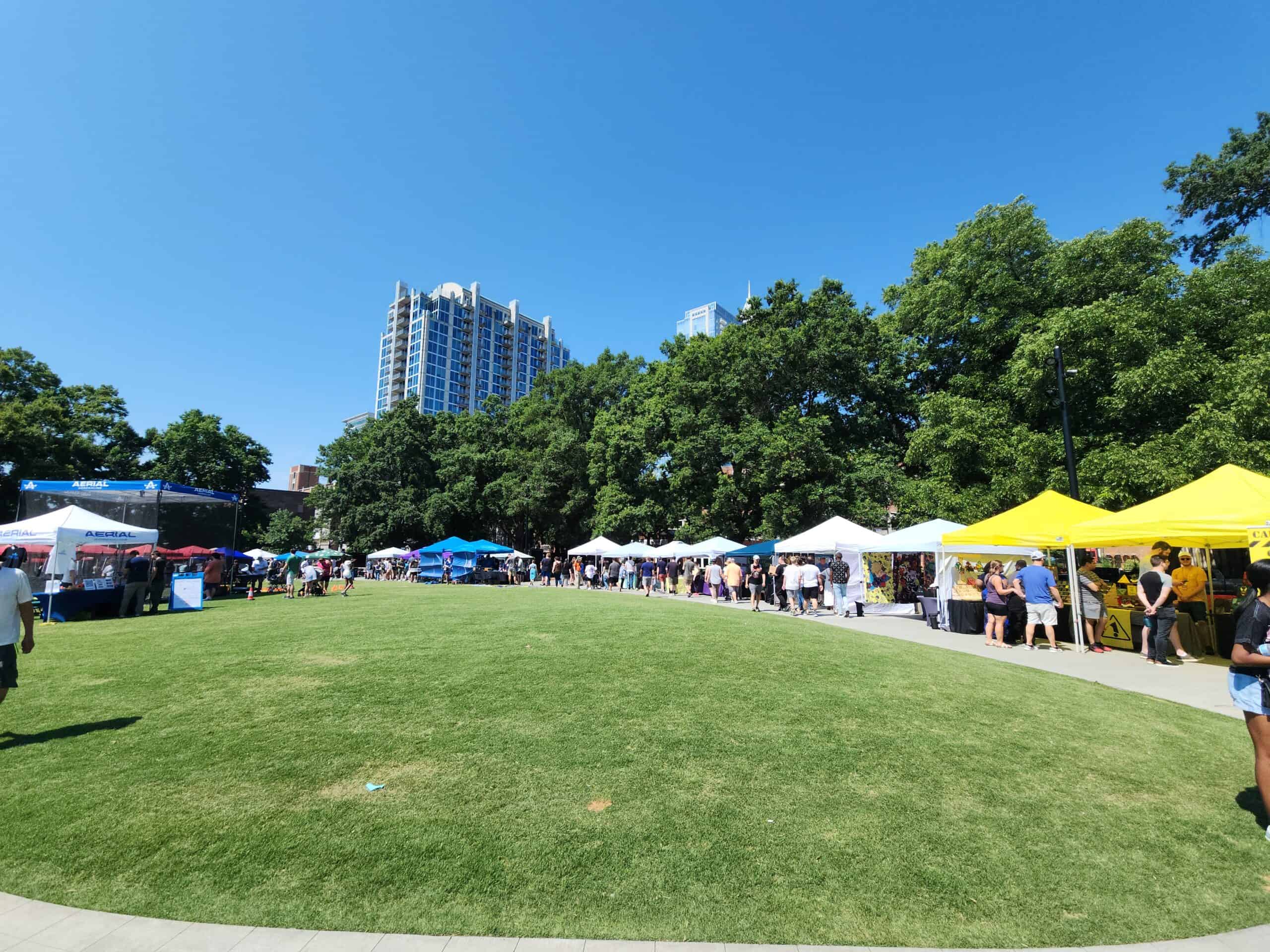 
A sunny outdoor event with numerous white and yellow tents lined up on one side of a large grassy area, surrounded by trees and high-rise buildings in the background. People are walking, browsing stalls, and enjoying the vibrant atmosphere under a clear blue sky.