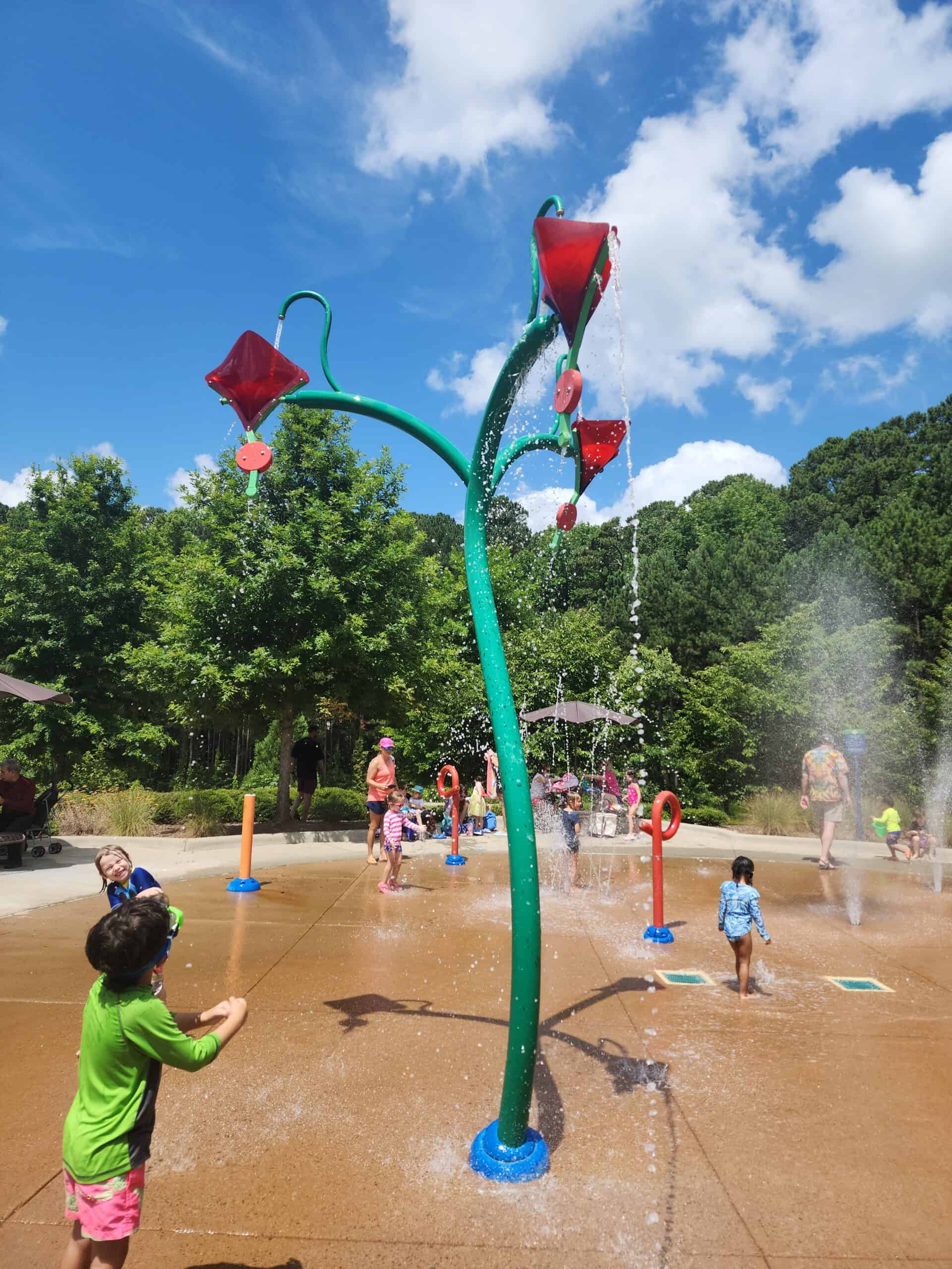 Children playing in a splash pad area with water features designed like colorful flowers, spraying water from overhead. The scene is lively with kids running through the water jets under a bright blue sky, surrounded by lush green trees. This is a fun and engaging activity for families looking for free things to do in Raleigh with kids.