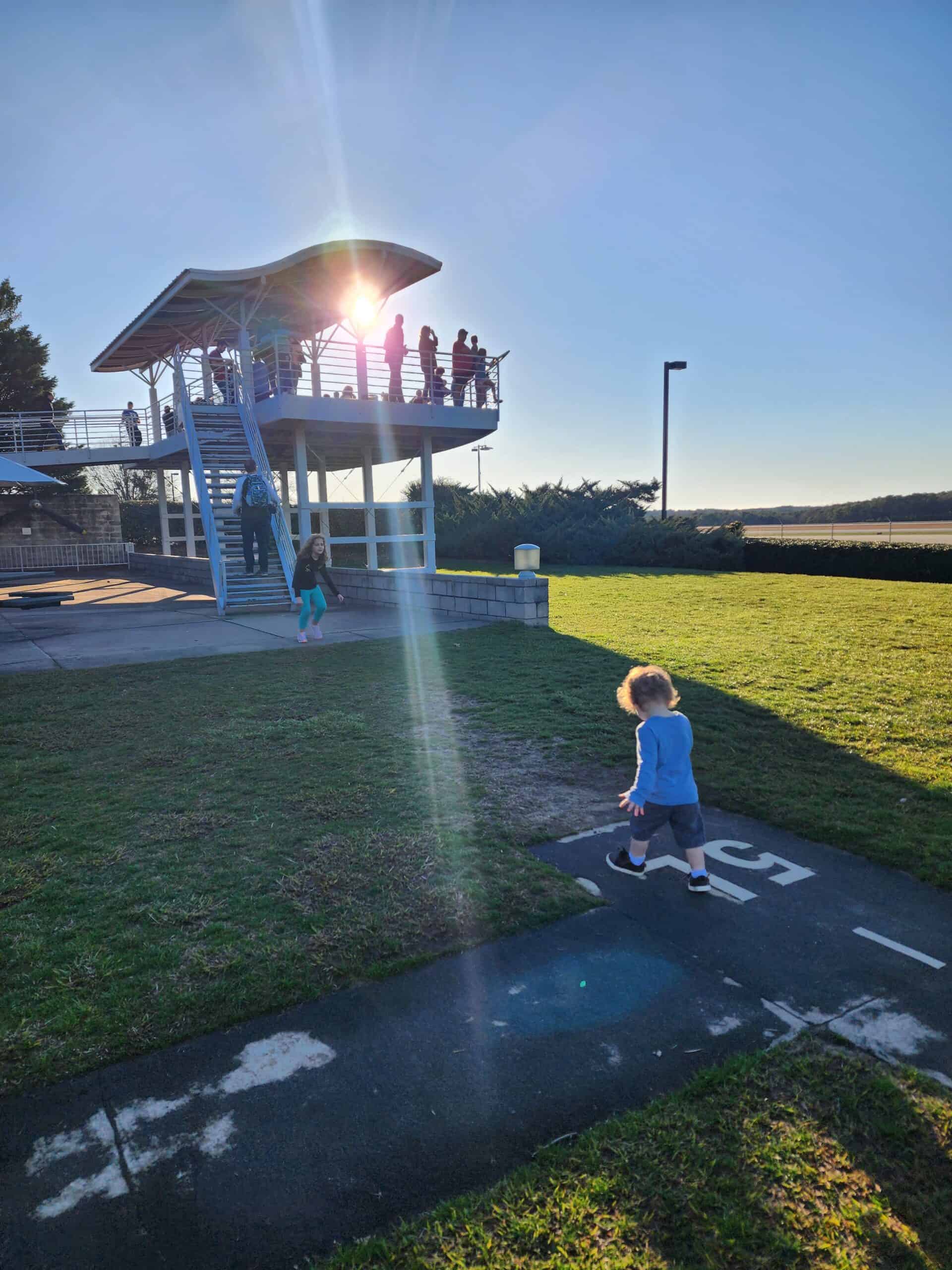 
Children playing near a viewing platform during sunset, with the sun shining through the structure. The platform is elevated, accessible by stairs, and has several people enjoying the view from the top. The area around is grassy with a clear, blue sky overhead, creating a peaceful and scenic environment.