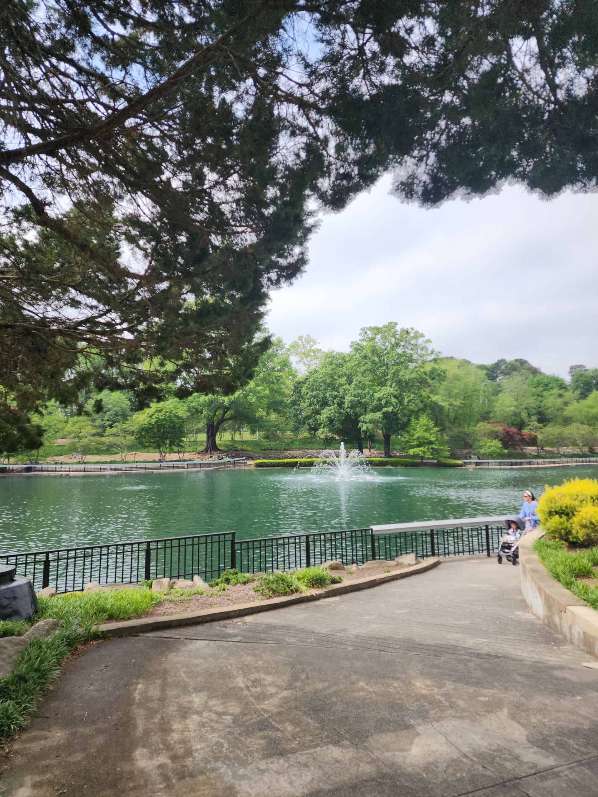 A serene lake scene featuring a fountain in the center, surrounded by lush greenery and trees. The image captures a walkway leading to the lake, bordered by black railings, with a person pushing a stroller along the path. The overhanging tree branches frame the view, creating a picturesque and tranquil atmosphere.