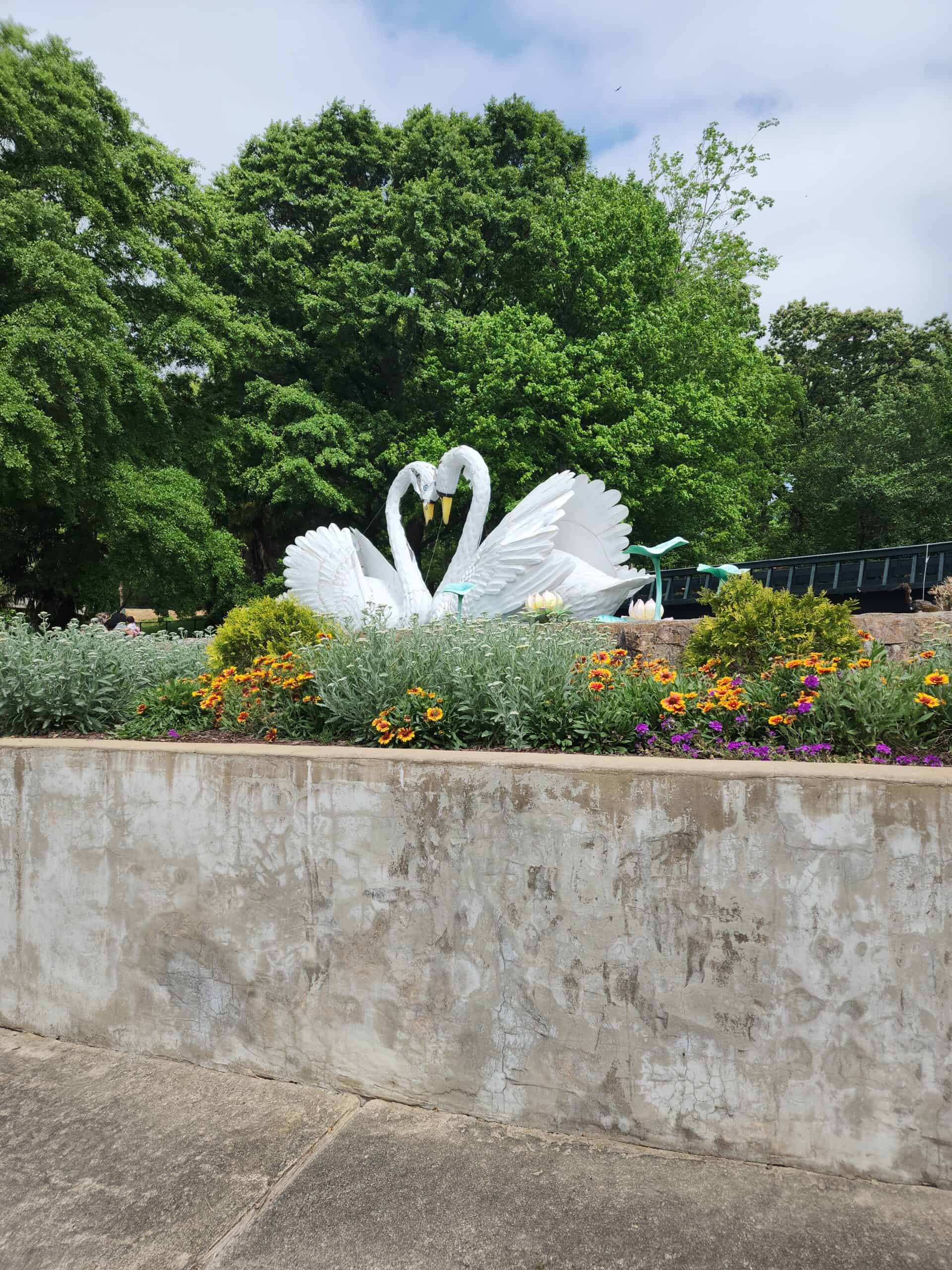 A concrete structure with vibrant flowers in front, featuring a sculpture of two large white swans with necks intertwined, forming a heart shape. The background is filled with lush green trees under a partly cloudy sky.