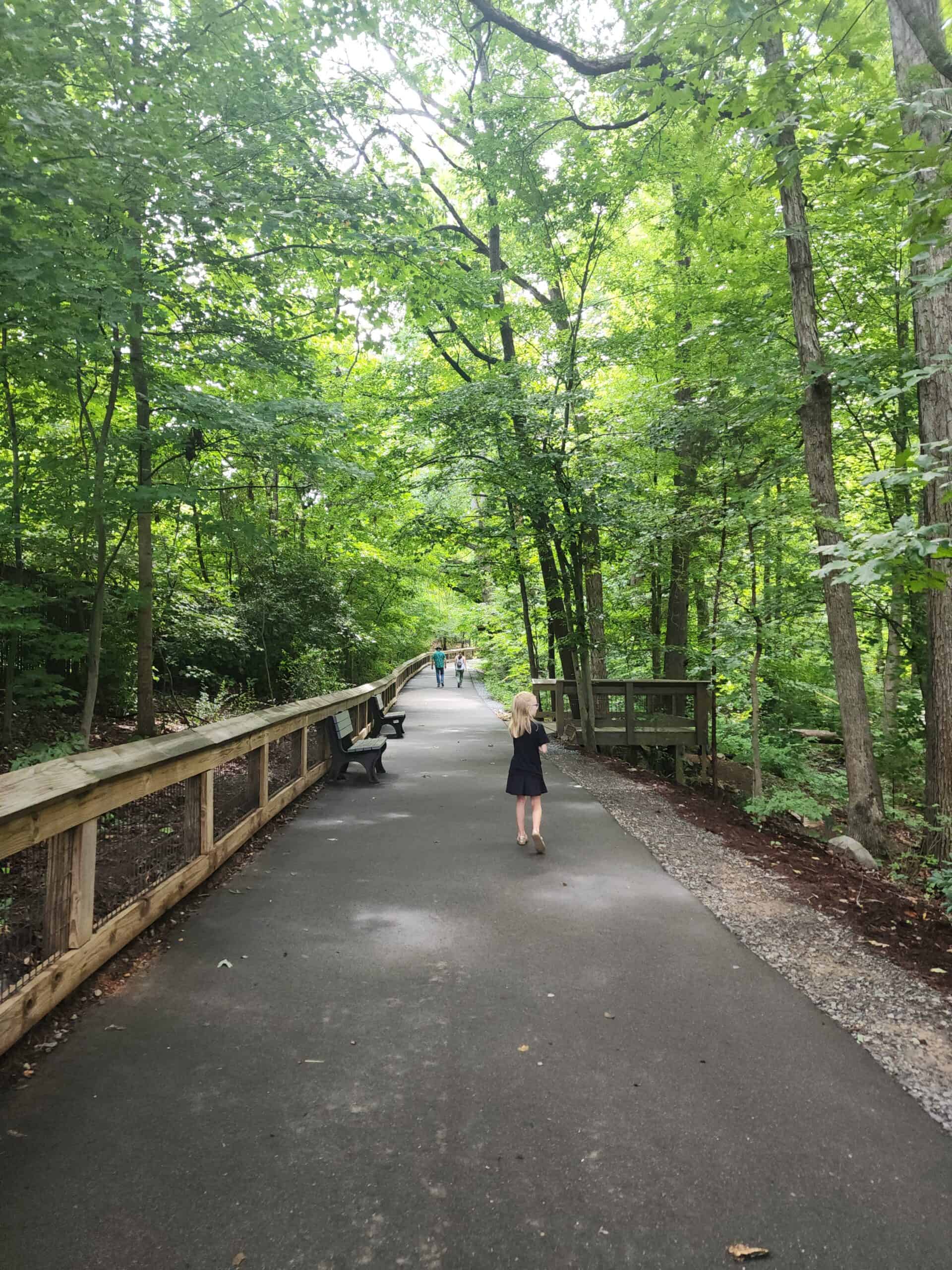 A young girl walks along a paved path surrounded by lush green trees in a forested area, with a wooden fence and benches lining the trail. Other visitors can be seen further down the path, enjoying the serene natural environment.