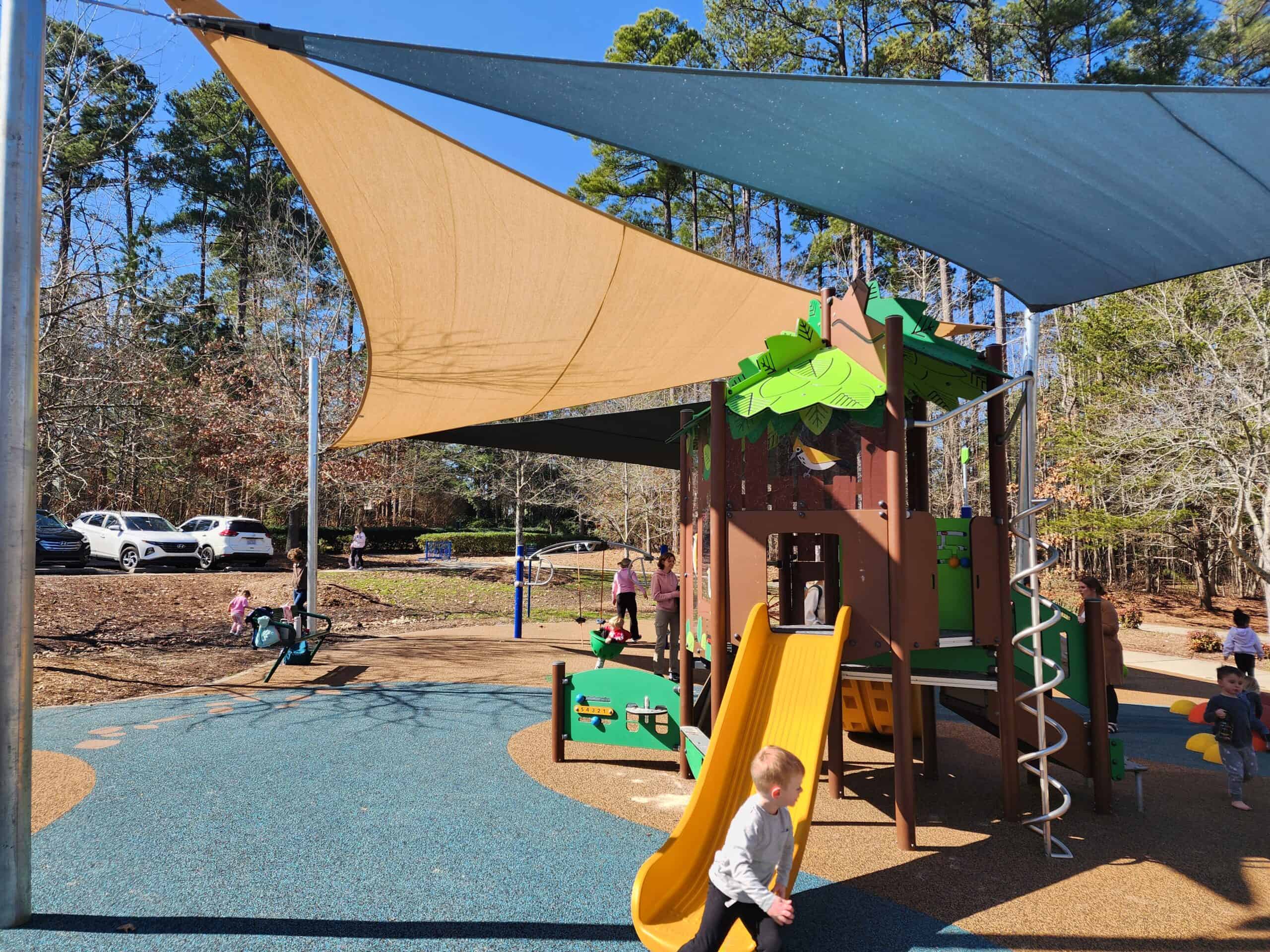 A children's playground with a nature-themed play structure featuring a yellow slide, green climbing elements, and a spiral ladder. The area is shaded by large triangular fabric canopies in tan and blue, providing protection from the sun. The playground surface is covered with blue and brown rubber flooring. Several children are playing, and a few adults are supervising in the background. Trees and parked cars are visible beyond the playground, indicating a wooded, suburban setting. This image highlights a shaded, well-designed play area ideal for young children