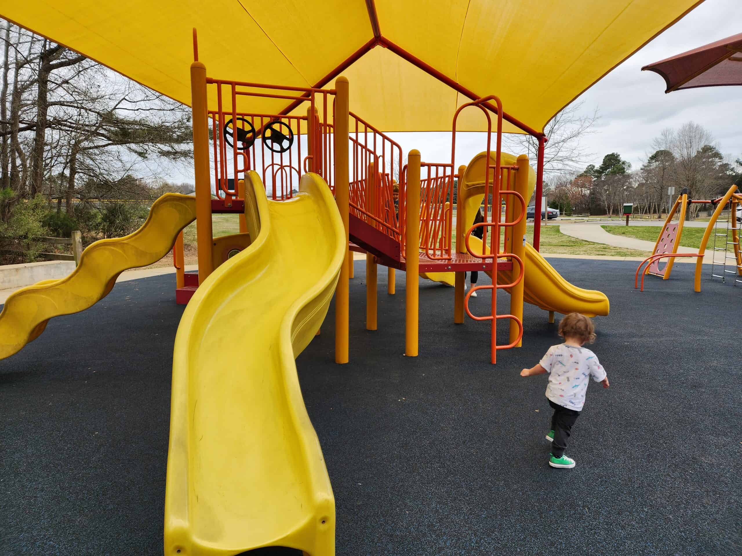 A playground with bright yellow and orange slides and climbing structures, set on a blue rubber safety surface. The area is shaded by a large yellow canopy overhead. A small child in a white shirt and green shoes walks across the playground, adding a sense of activity. The playground is surrounded by open space and trees, with a cloudy sky overhead, suggesting a cool and comfortable environment for play.