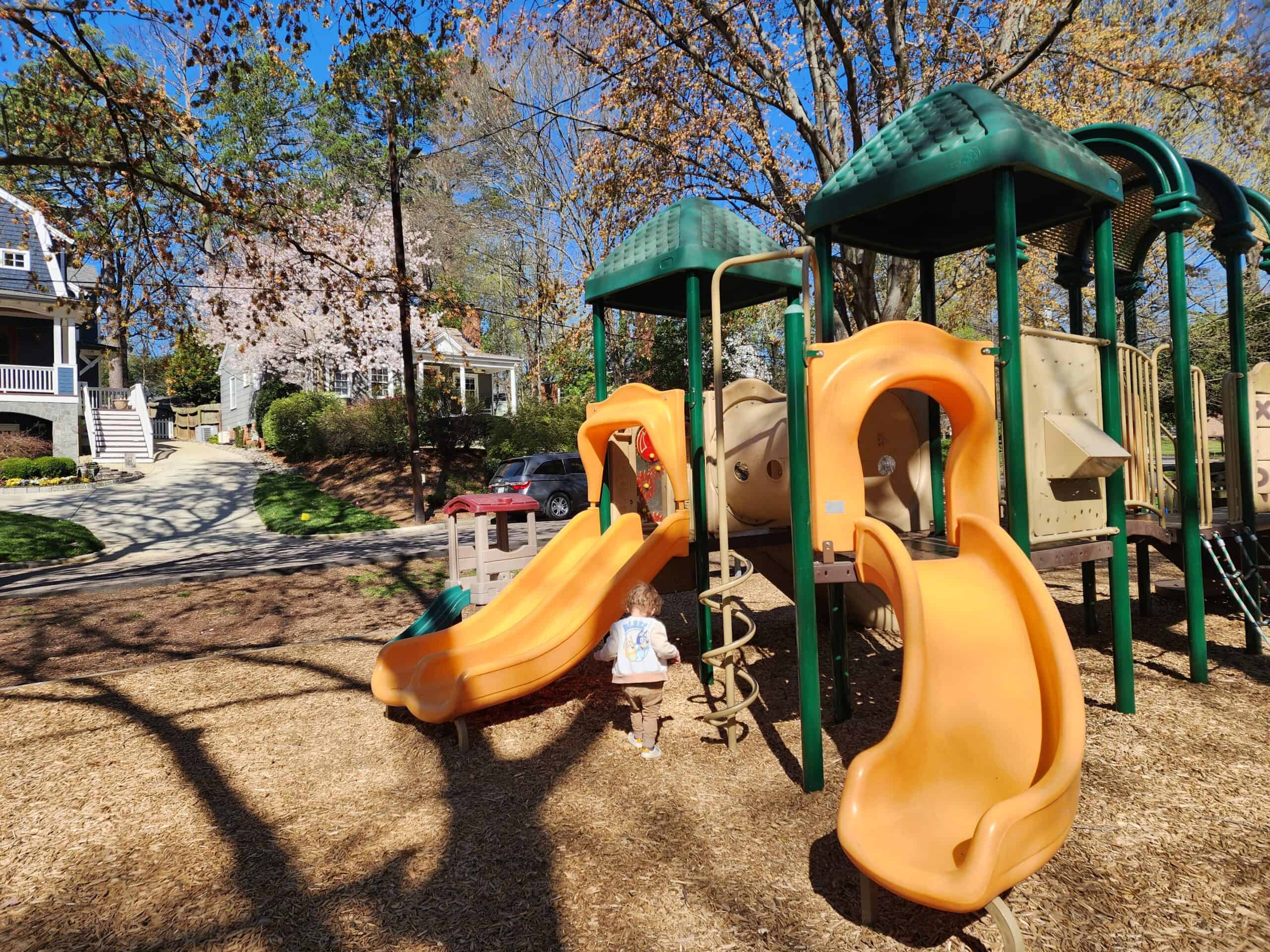 A neighborhood playground with green-roofed play structures and orange slides, surrounded by wood chip ground cover. A young child is seen playing near the base of the slides. The playground is set within a residential area, with houses and blooming trees visible in the background. The scene is bright and sunny, with shadows of trees cast on the ground, suggesting a pleasant day for outdoor play.