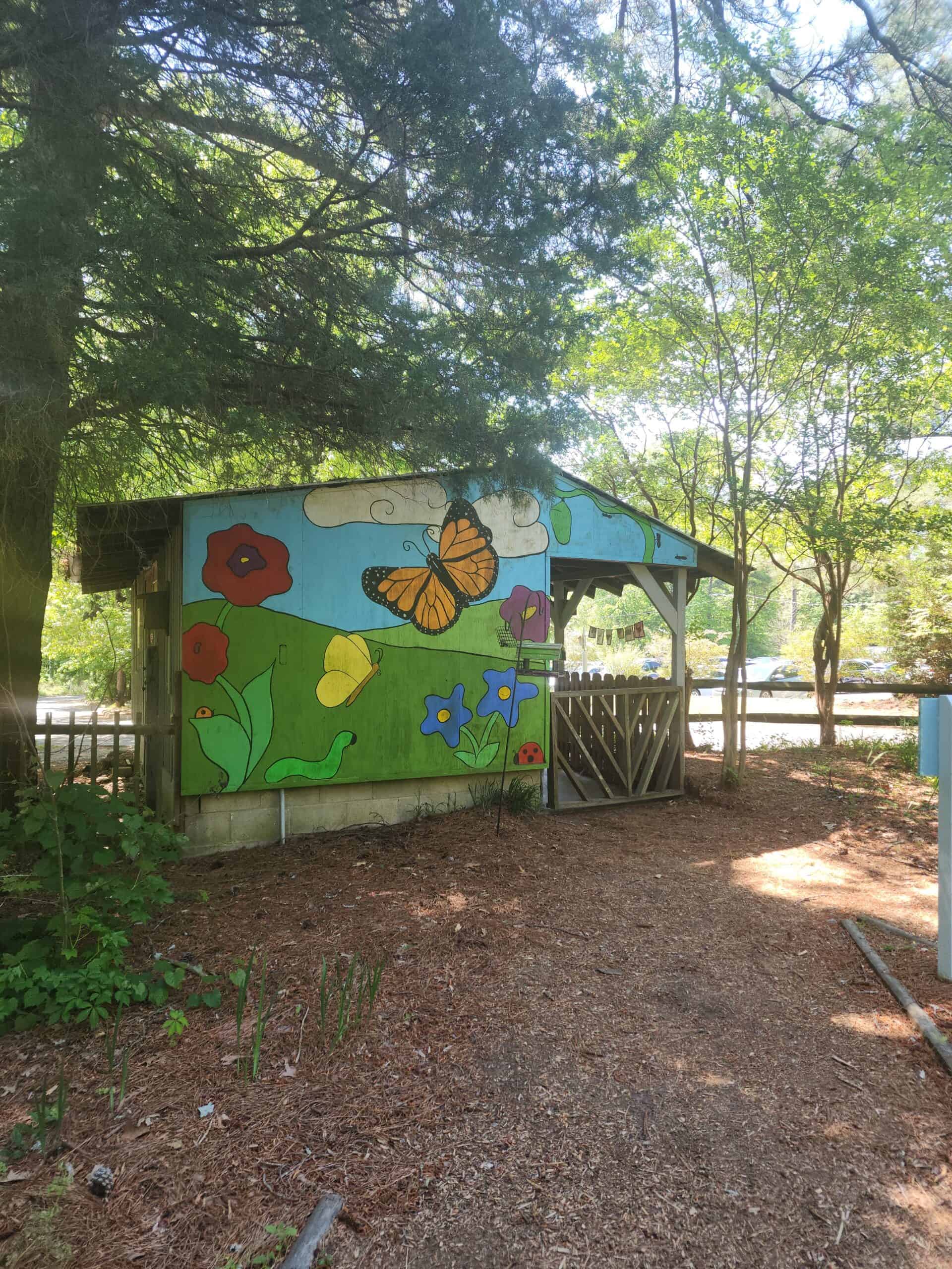 A small wooden structure surrounded by trees, featuring a brightly painted mural on its side. The mural depicts a colorful scene with a large orange butterfly, red and blue flowers, and green leaves under a partly cloudy sky. The structure sits along a dirt path, with shade provided by the surrounding trees. The overall setting suggests a peaceful, natural environment, possibly part of a park or garden area.