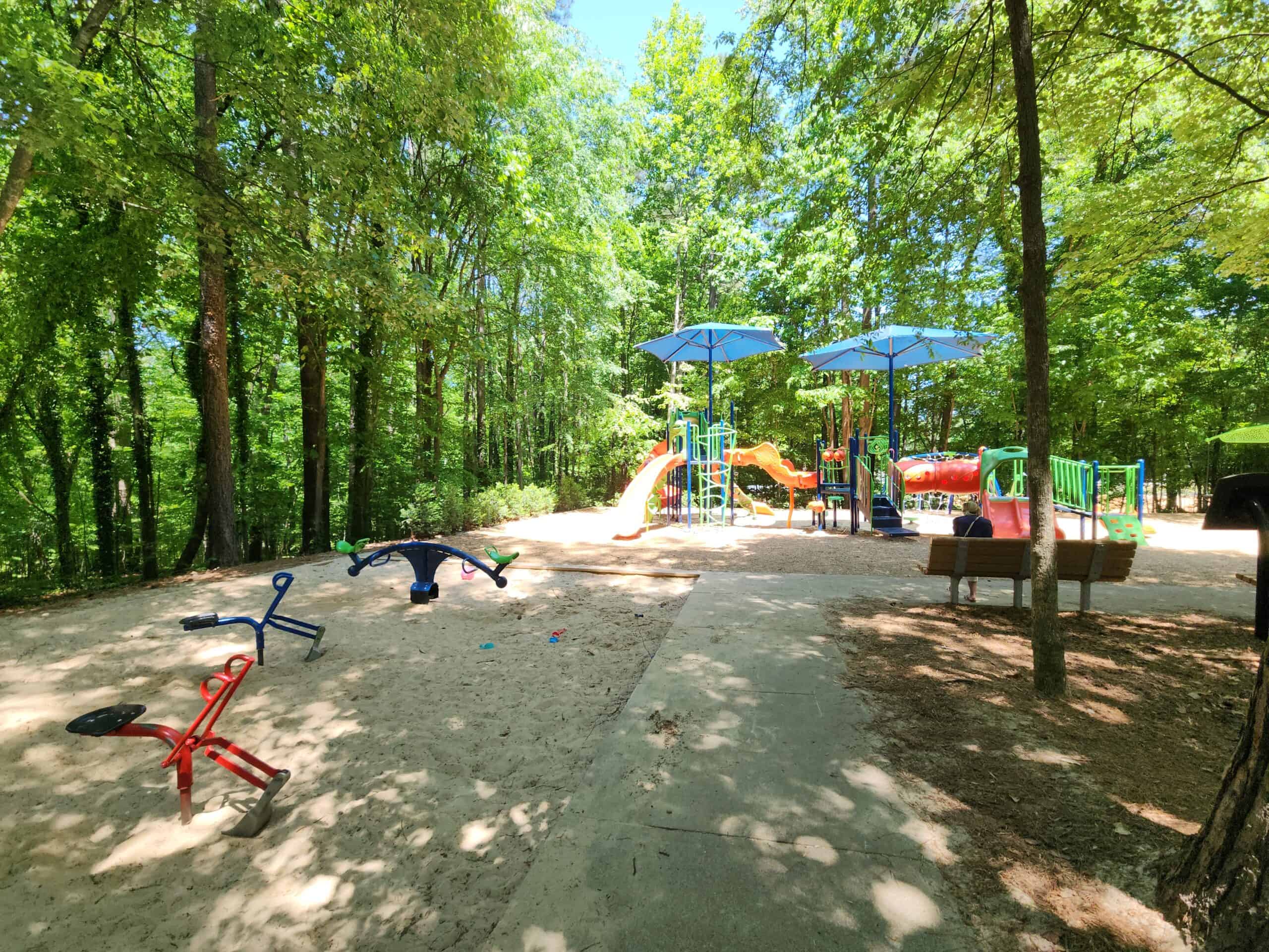 A shaded playground set within a wooded area, featuring colorful slides and climbing structures under large blue umbrellas. The ground is covered in sand, with a few seesaws scattered in the play area. A wooden bench is positioned nearby, with a person seated on it. The surrounding trees provide ample shade, creating a cool and serene environment for children to play in. This image highlights a peaceful, shaded playground surrounded by nature.