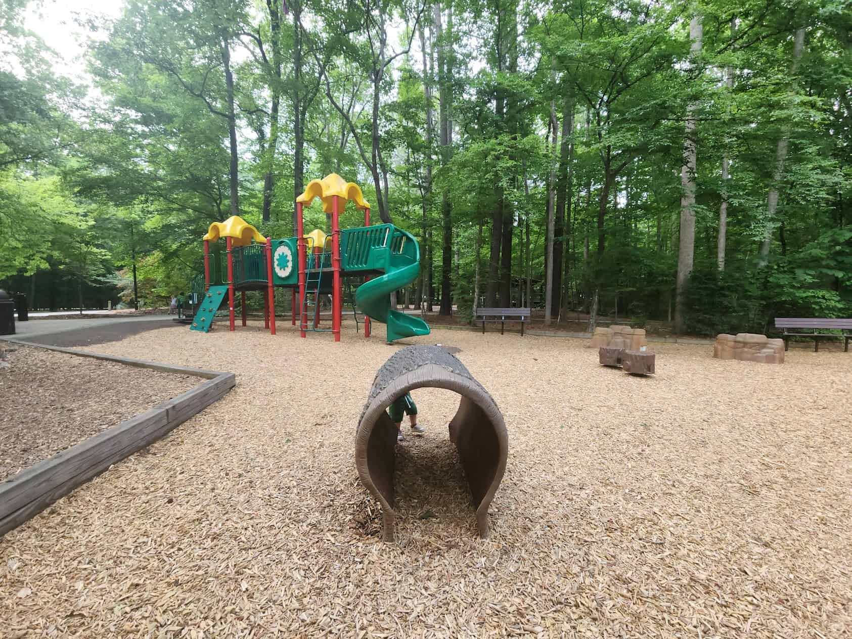 A playground set in a wooded area features bright green and yellow play structures, including slides, climbing walls, and tunnels. The ground is covered with wood chips, and there are benches and natural-looking log seats nearby. A small log tunnel is in the foreground, adding to the rustic and nature-inspired design of the playground. The surrounding trees provide ample shade, creating a peaceful and natural atmosphere.