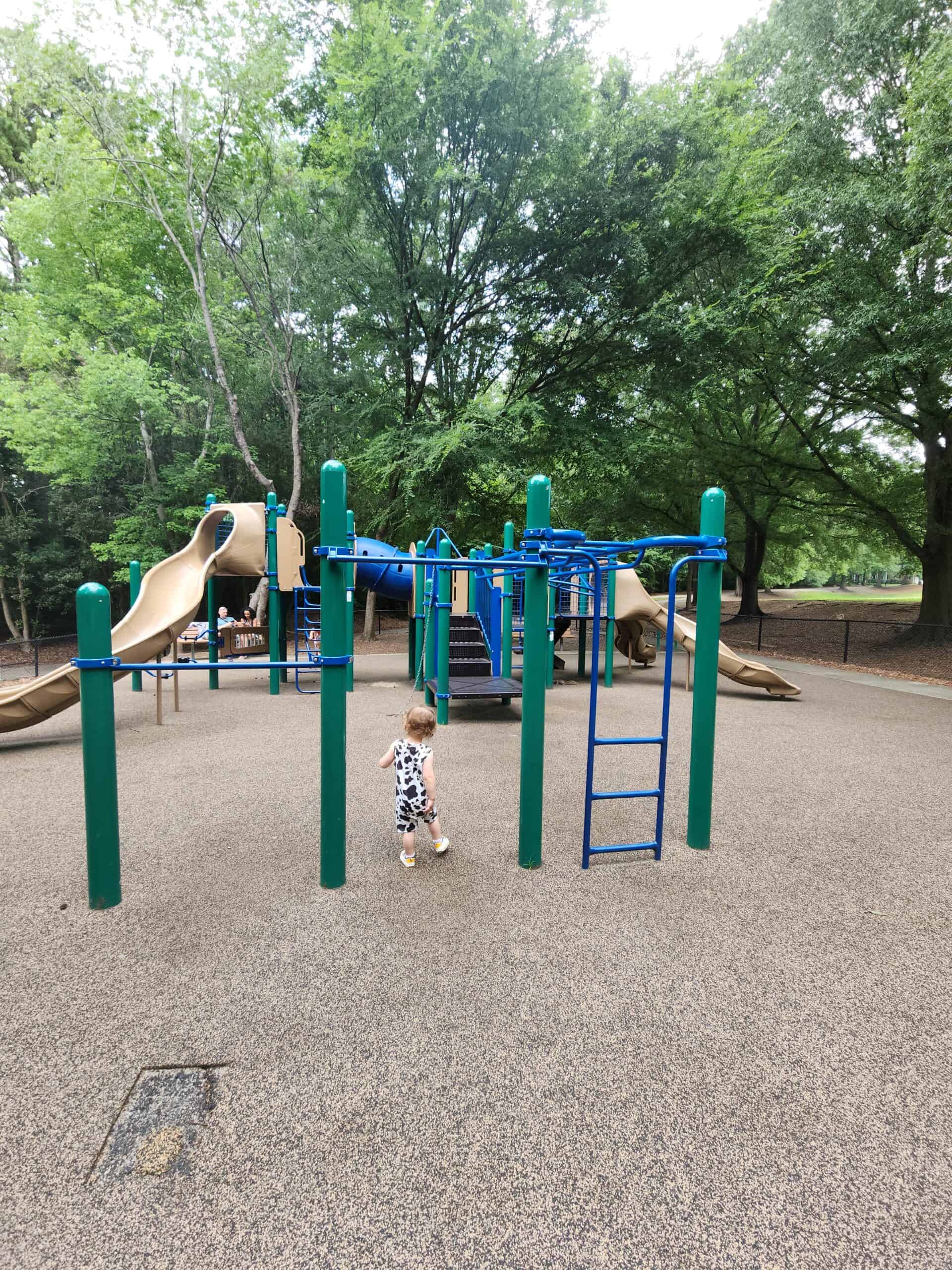 A playground with green and blue climbing structures and slides is set against a backdrop of tall, leafy trees that provide ample shade. The ground is covered with a rubber safety surface. A small child dressed in a cow-patterned outfit is walking across the playground, adding a sense of scale and activity. The scene is quiet and surrounded by nature, offering a peaceful environment for play.