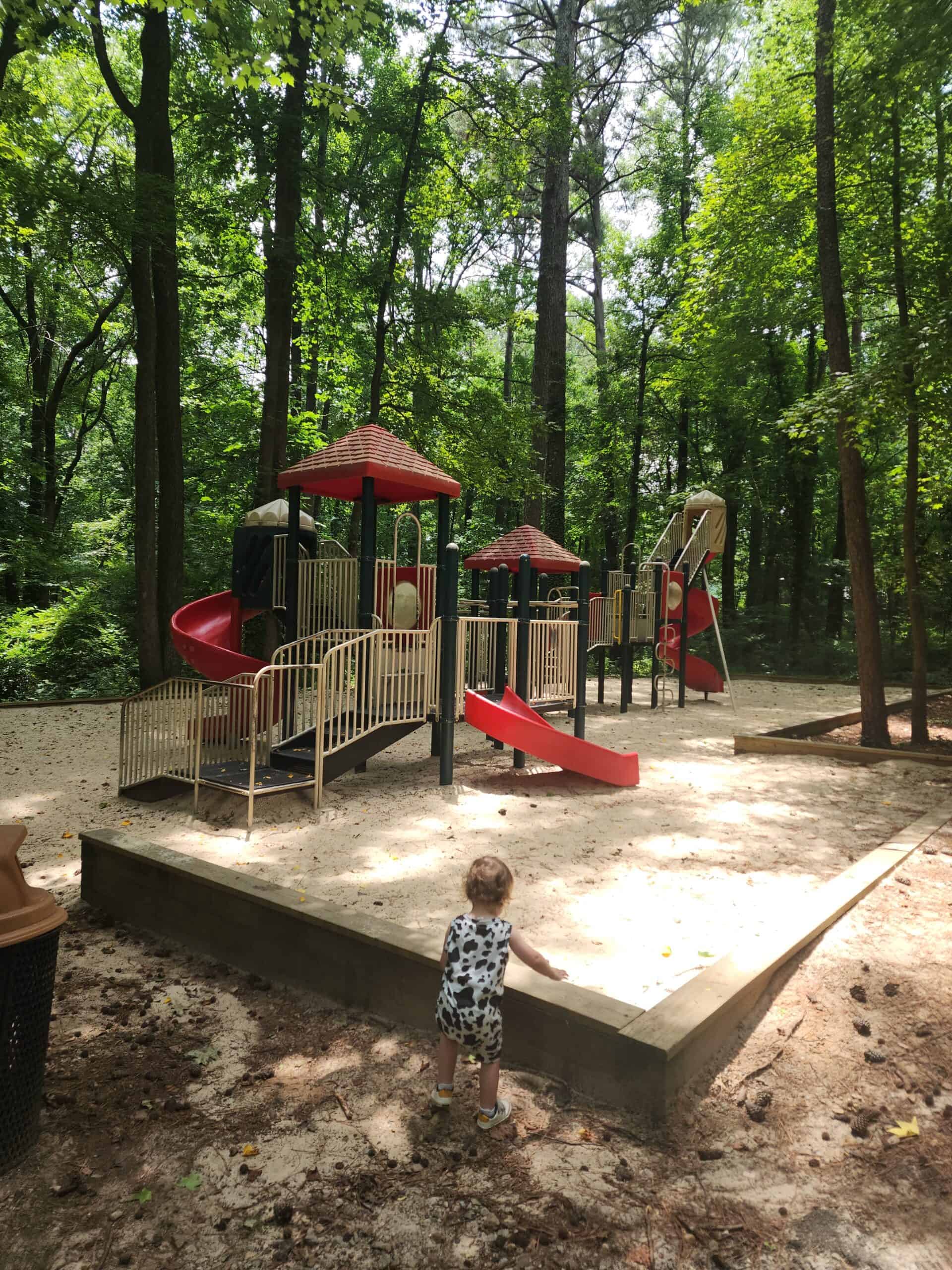 A shaded playground nestled in a wooded area, featuring red-roofed play structures with slides and climbing elements. The ground is covered with sand, and a small child in a cow-patterned outfit is standing near the edge of a raised sandbox area, facing the playground. Tall trees surround the play area, providing a natural and serene environment for outdoor play. The scene is quiet, with dappled sunlight filtering through the trees.