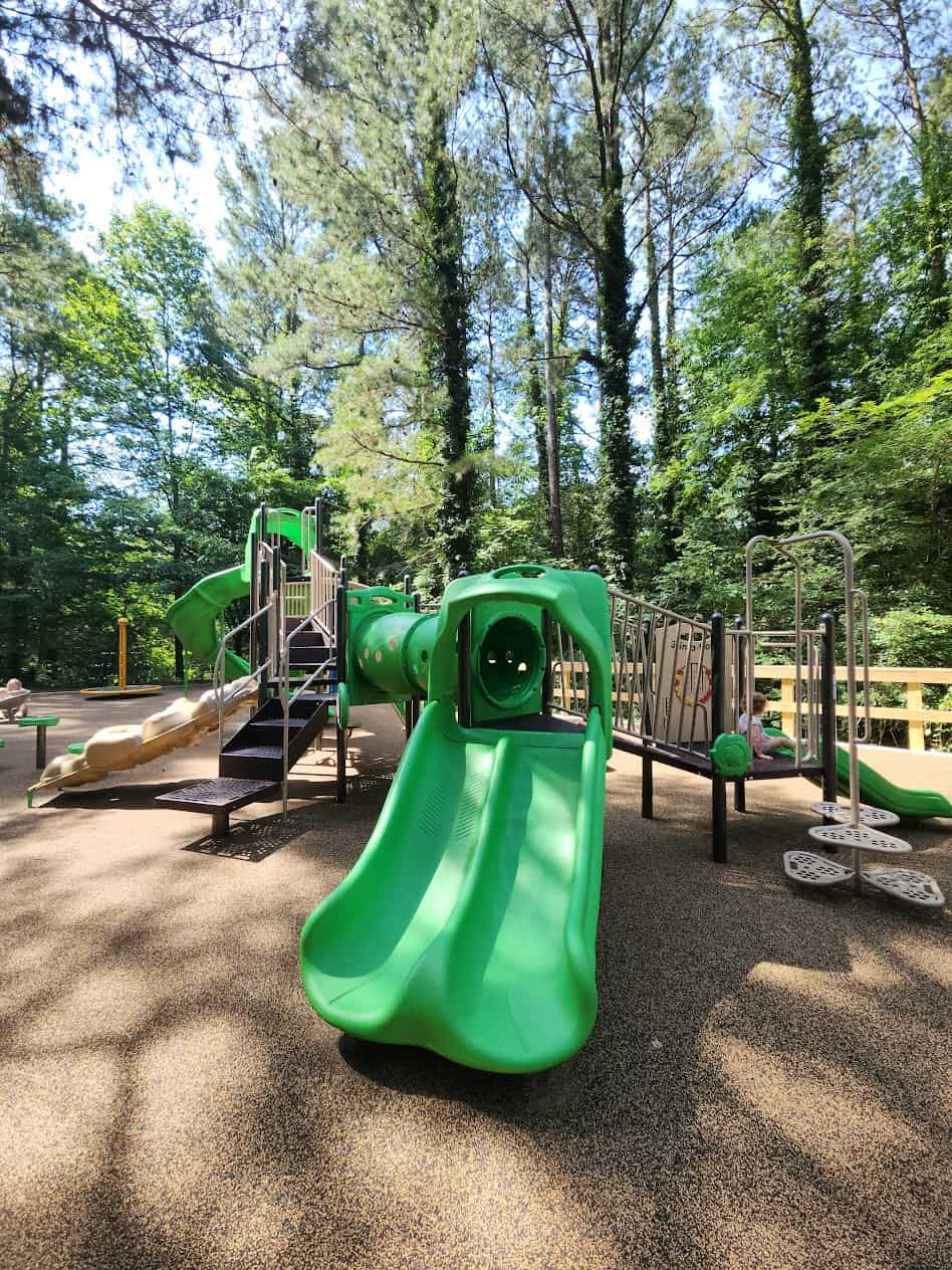 
A vibrant playground set amidst a forest features green play equipment, including slides, climbing structures, and tunnels. The playground is surrounded by tall trees, providing ample shade and a natural backdrop. The ground is covered with a soft, rubberized surface, ensuring safety for children playing. The atmosphere is tranquil, with dappled sunlight filtering through the trees, creating a blend of light and shadow across the playground