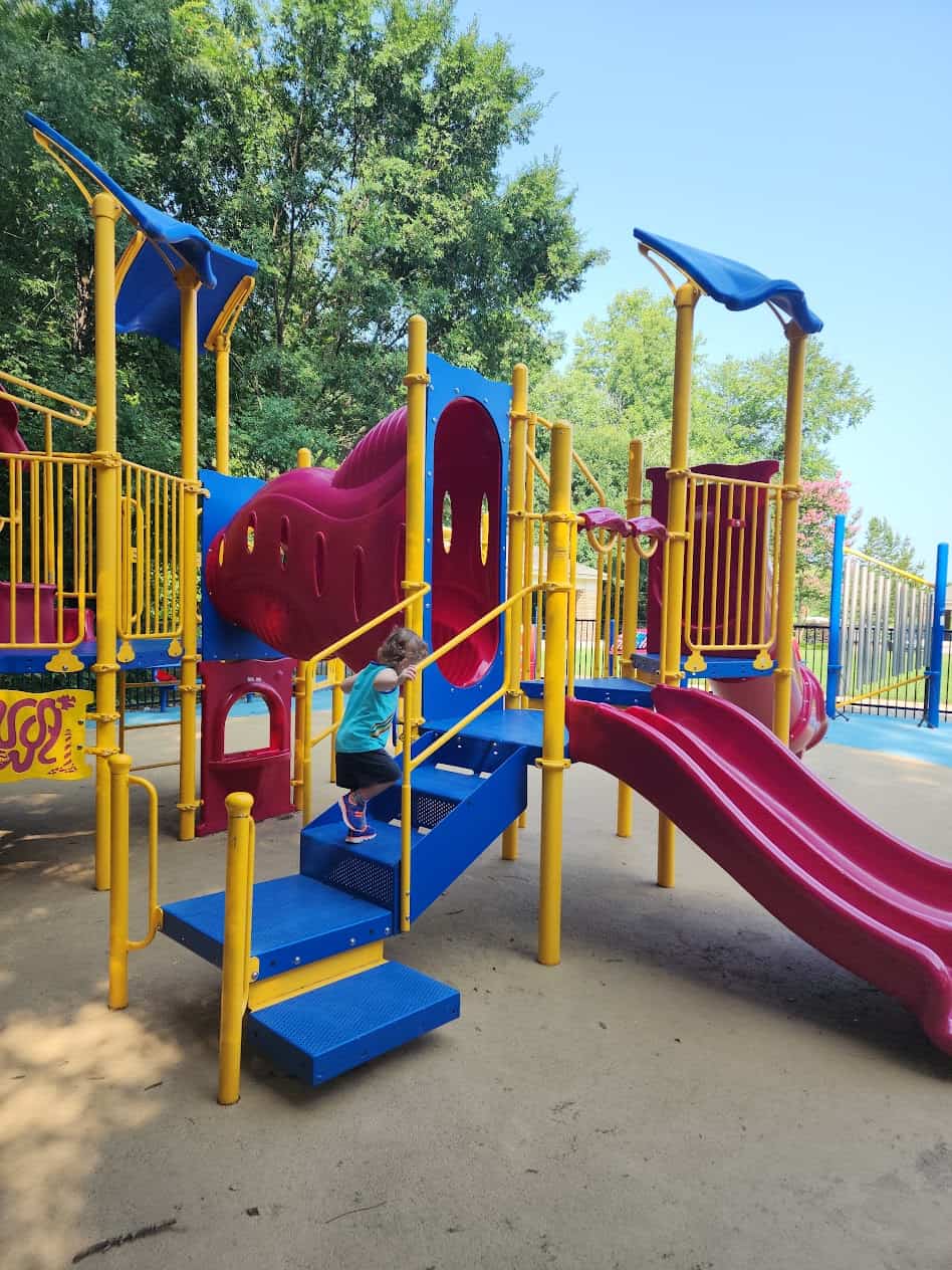 A young child is climbing the stairs of a brightly colored playground structure. The playground features yellow, red, and blue equipment, including slides and tunnels, with a few blue canopies overhead for shade. The area is surrounded by green trees, suggesting it is located in a park or a well-shaded outdoor play area. The day appears to be sunny and clear.