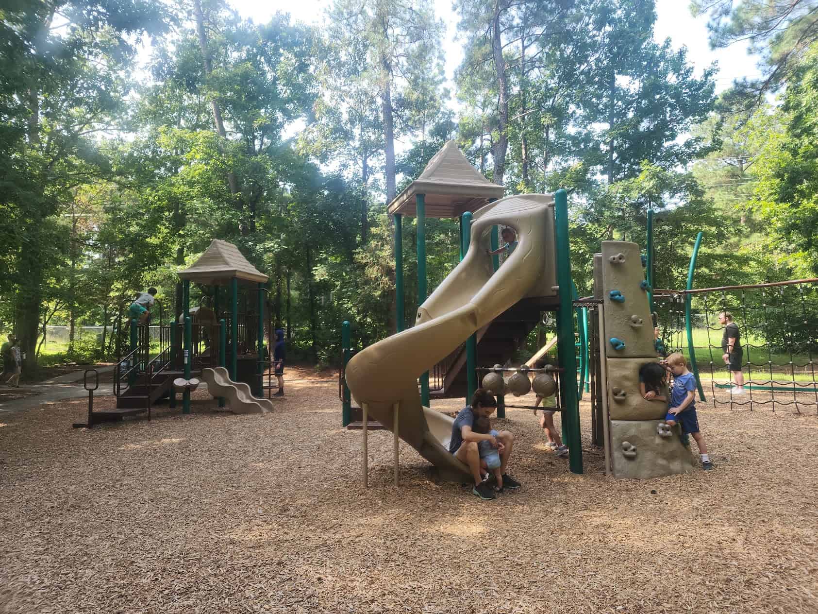 Children are playing on a playground surrounded by tall trees, giving the area a shaded, forest-like atmosphere. The playground equipment includes slides, climbing structures, and a small rock wall. The ground is covered with wood chips, and the setting is likely a park with a natural, wooded environment. A few adults are present, supervising the children as they play. The day appears to be warm and sunny.