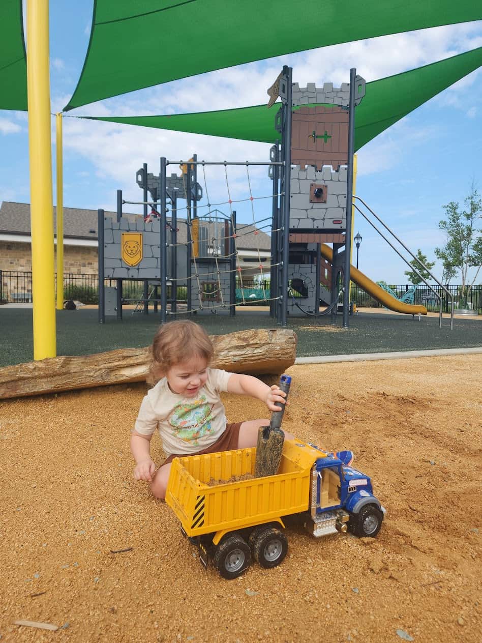 A young child is playing in a sandbox with a toy dump truck. The child is scooping sand with a small shovel into the yellow and blue dump truck. In the background, there is a playground structure resembling a castle with a climbing net and slide, partially shaded by green canopies. The setting appears to be an outdoor playground on a sunny day.