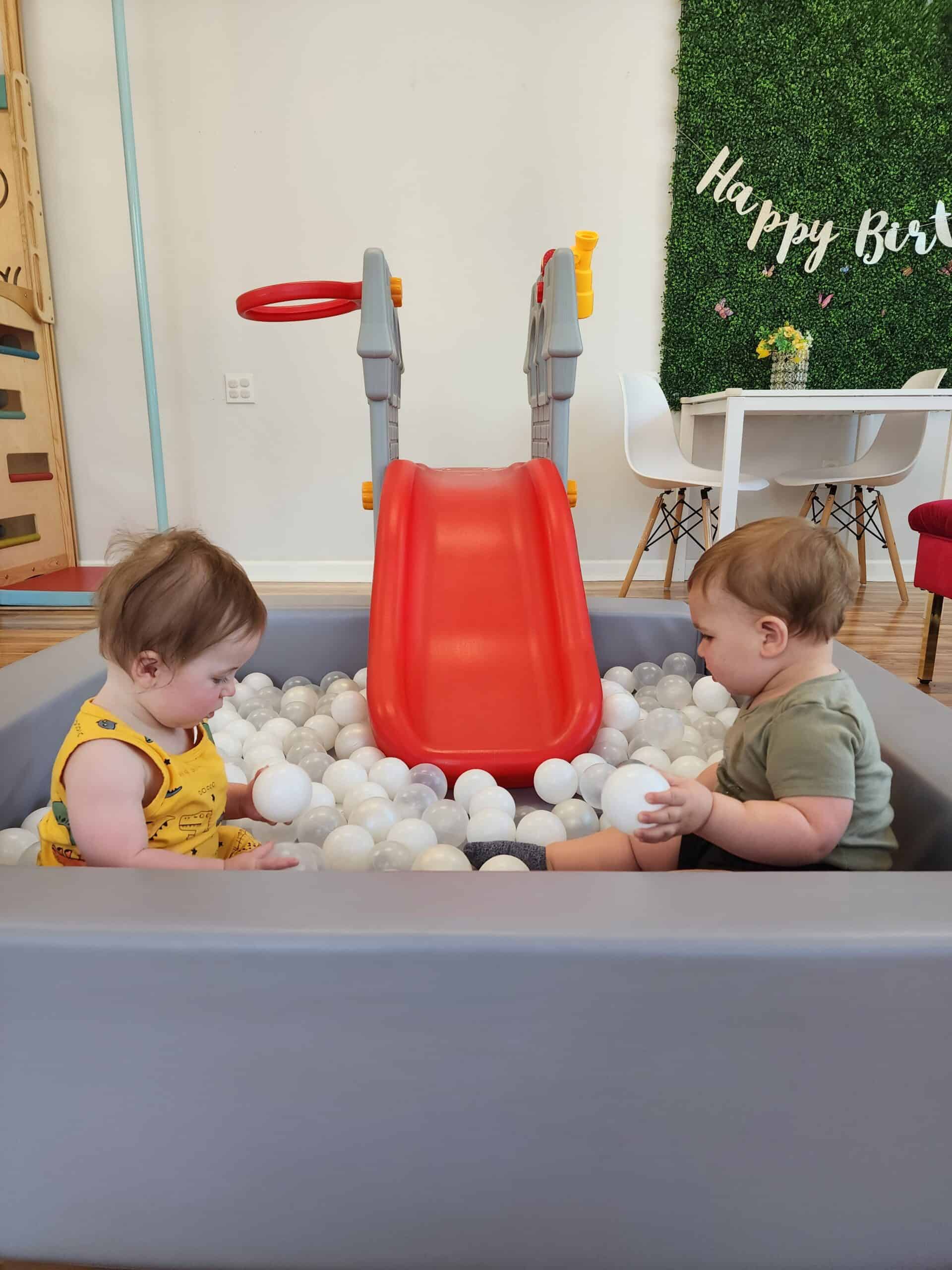 This image shows two young children playing in a small ball pit filled with white and translucent balls. A bright red plastic slide leads into the pit, adding to the playful setup. In the background, a green wall with the words "Happy Birthday" suggests this area might be part of a celebration or party venue, complemented by a small table with yellow flowers.