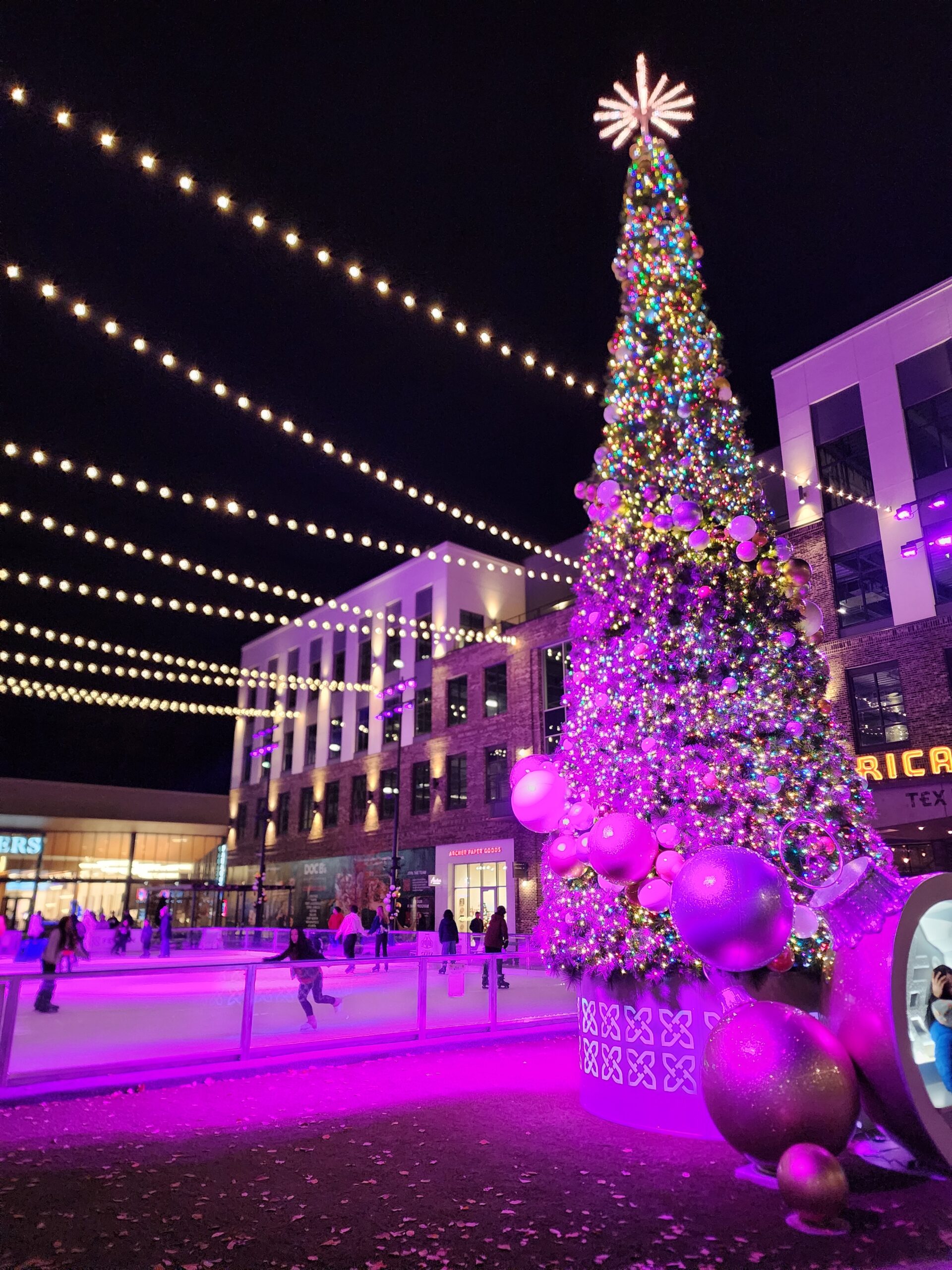 The image shows a festive outdoor scene featuring a large, brightly lit Christmas tree adorned with colorful ornaments and topped with a glowing star-like decoration. Beneath strings of warm fairy lights, people are skating on an ice rink illuminated in soft purple hues, creating a vibrant holiday ambiance. The setting appears to be in front of modern buildings, adding a lively, urban charm to the celebration.