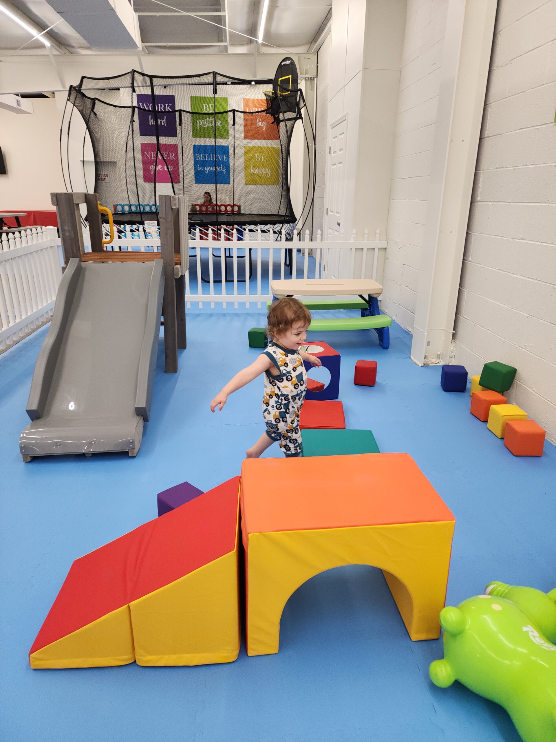 This image depicts a bright indoor play area featuring a soft obstacle course with colorful foam blocks, ramps, and tunnels. A toddler is actively playing, surrounded by vibrant equipment, including a small gray slide and a green inflatable toy. In the background, a trampoline with motivational signs, such as "Believe in yourself" and "Be happy," adds a positive and playful atmosphere.