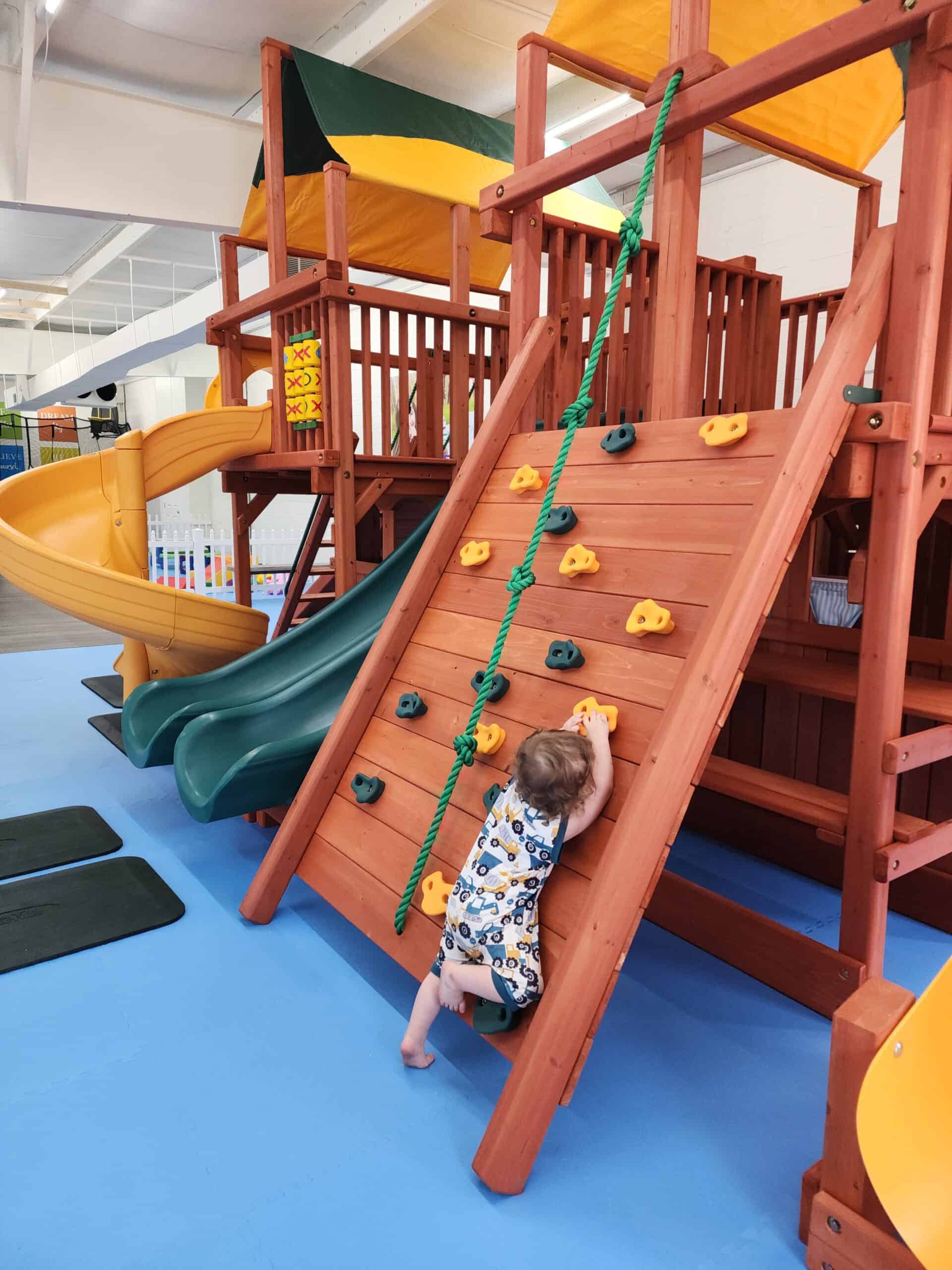 This image features a wooden indoor playground structure with a child climbing a small rock wall equipped with colorful grips and a green rope for support. The setup includes dual green and yellow slides on the side, topped with yellow and green canopies, creating a cozy play fort vibe. The soft blue flooring ensures safety, making the area perfect for adventurous play.