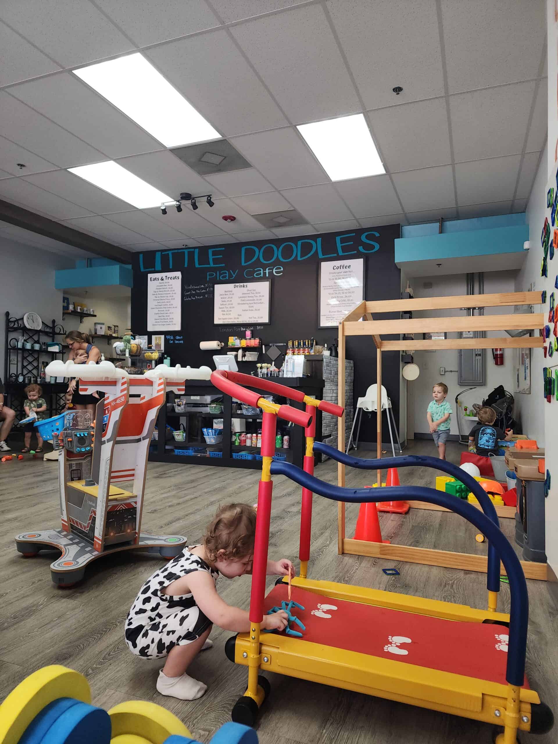 This image captures a cozy, child-friendly space at "Little Doodles Play Cafe," featuring a play area with colorful toys and play equipment. A toddler is seen interacting with a small red and yellow toy treadmill, while another child plays in the background. The cafe section in the back displays a blackboard menu with options for coffee, drinks, and snacks, emphasizing the venue's dual function as a play zone and relaxation spot for parents.