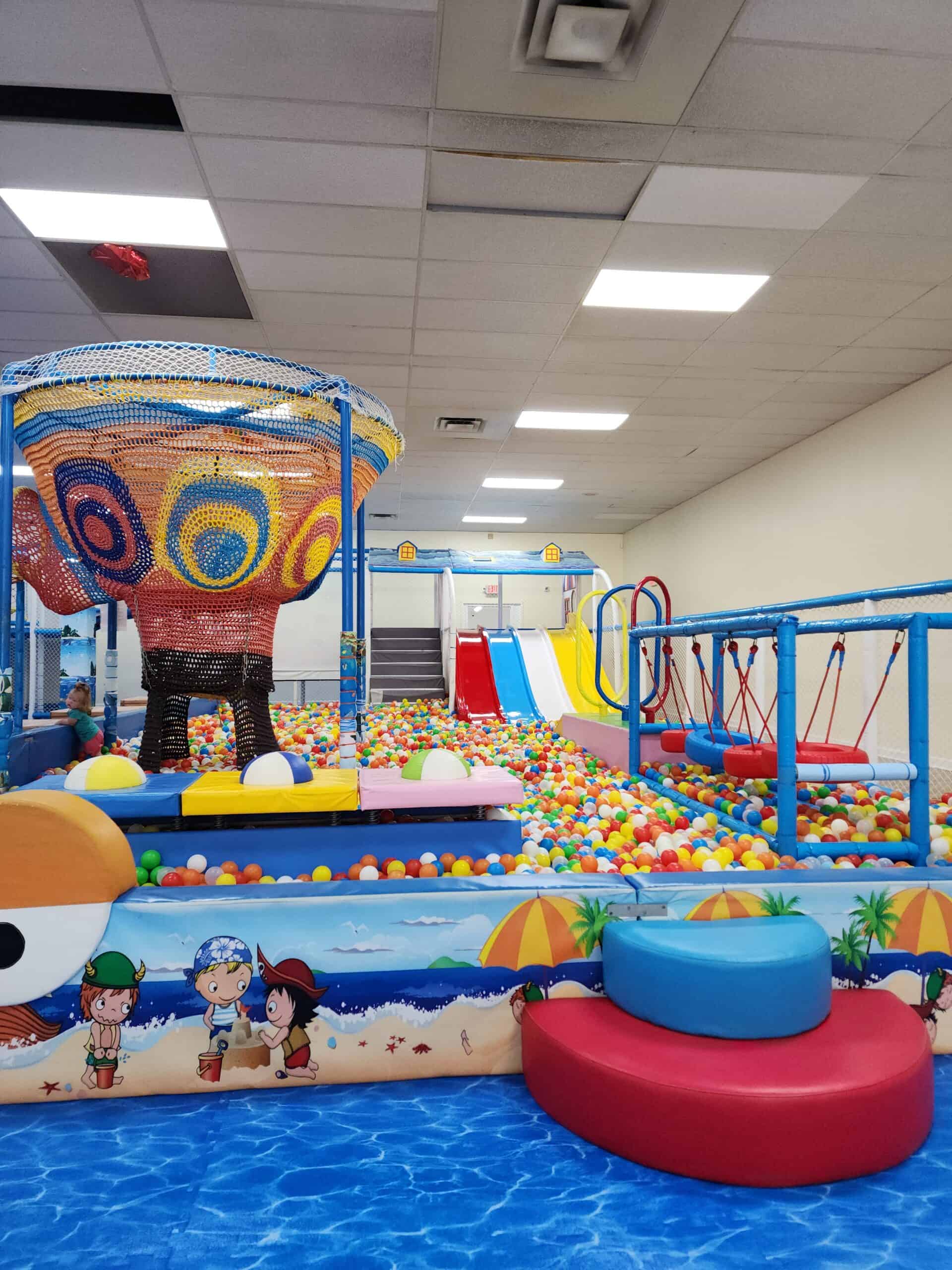 This image showcases a lively indoor playground filled with a large ball pit, colorful slides, and swings. A vibrant woven climbing structure with intricate patterns in red, yellow, and blue sits prominently at the front, inviting kids to explore. The play area features a beach-themed design with cartoon illustrations along the walls, enhancing the playful atmosphere. Splashes of primary colors across the equipment and the abundance of balls create a fun and energetic space for children.