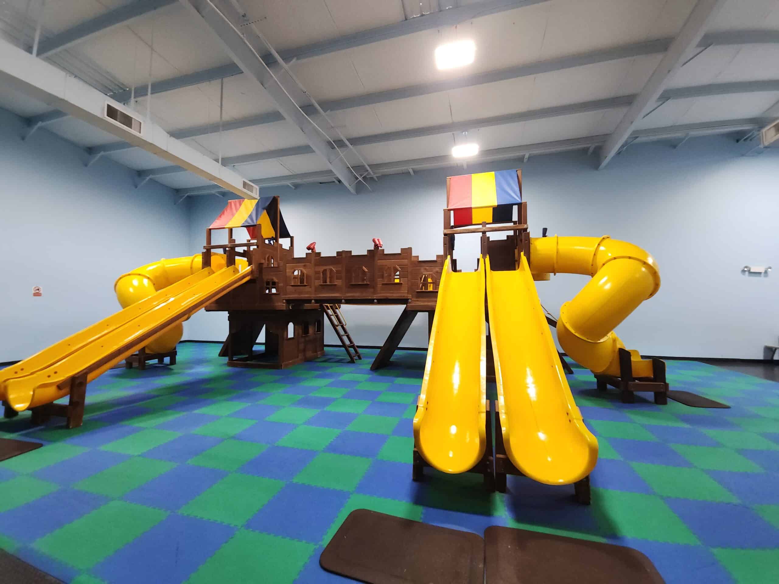 This image shows an indoor playground featuring a castle-themed wooden structure with vibrant yellow slides and enclosed tube slides. The play area has colorful blue and green padded flooring, enhancing safety and comfort for children. Two small tents in primary colors sit atop the structure, adding a whimsical touch to the castle-like design.