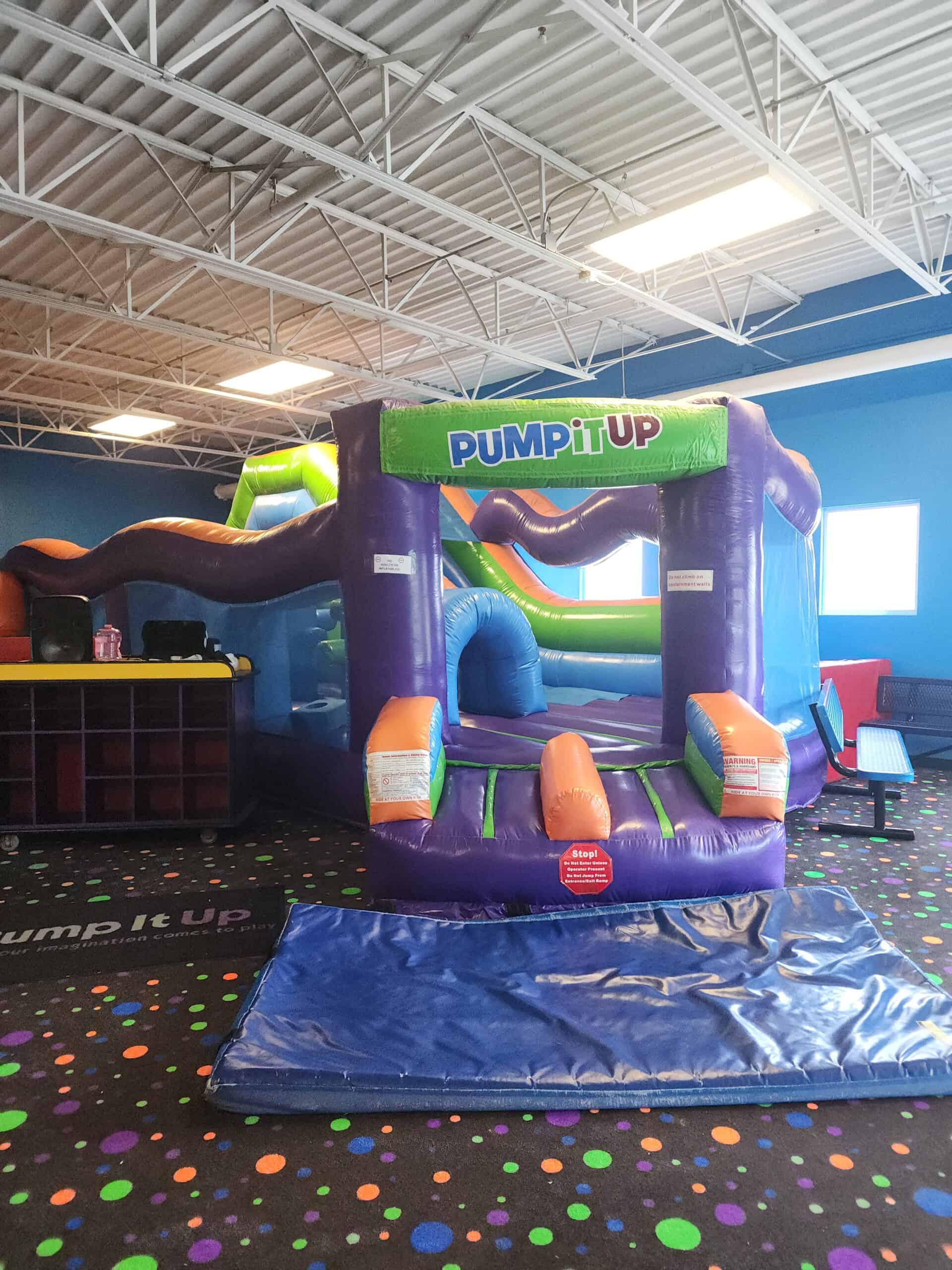 This image shows an indoor inflatable play structure from "Pump It Up," featuring colorful sections in purple, orange, and green. The inflatable is surrounded by a polka-dotted carpet with vibrant colors, suggesting a fun and playful environment designed for kids' activities. A padded mat lies at the entrance, emphasizing safety