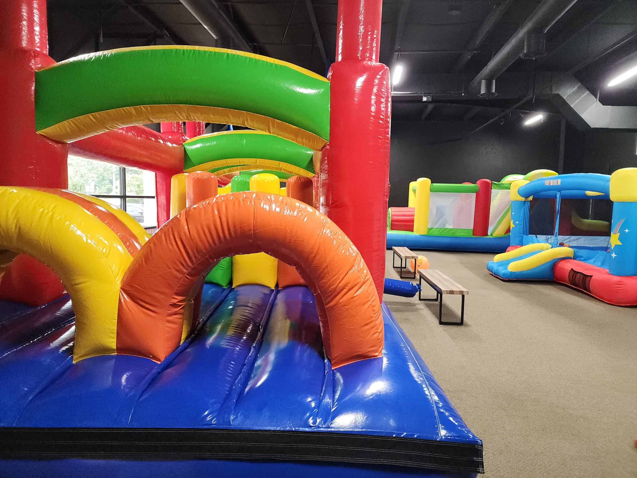 This image showcases an indoor inflatable play area with multiple bounce houses in bright, primary colors like red, green, yellow, and blue. The foreground features a large inflatable obstacle course with arches and padded sections, inviting active play. In the background, more inflatable structures are visible, including a smaller bounce house with a slide, creating a lively and energetic environment perfect for kids to run, jump, and explore.