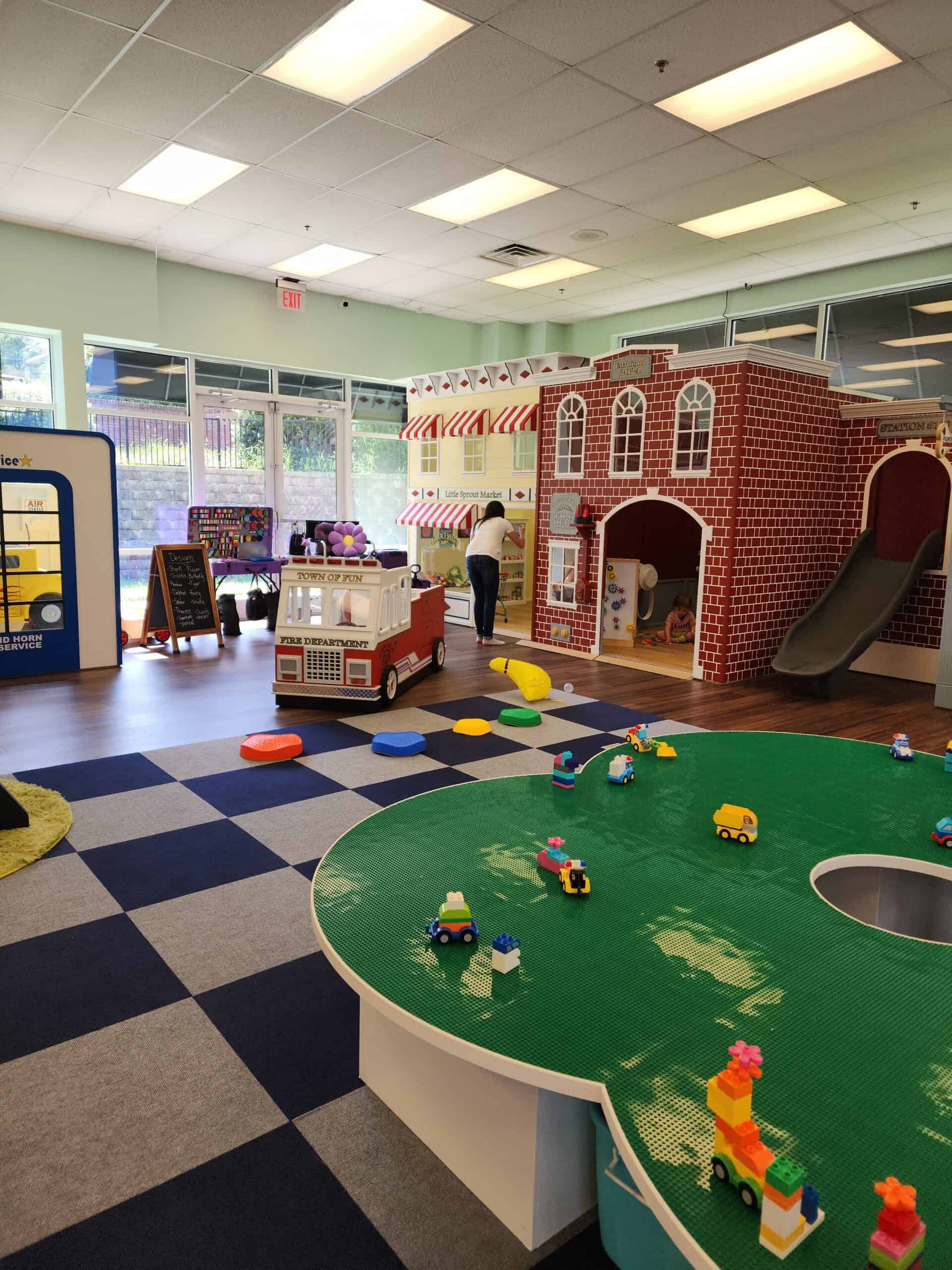 A wide-angle view of Town of Fun indoor playground, showcasing a Lego play table with small vehicles and block creations in the foreground. In the background, a fire truck prop, Little Sprout Market storefront, and a red-brick fire station with a slide contribute to the imaginative town theme. Bright natural light fills the space, and colorful play mats, props, and activity stations are scattered throughout the room