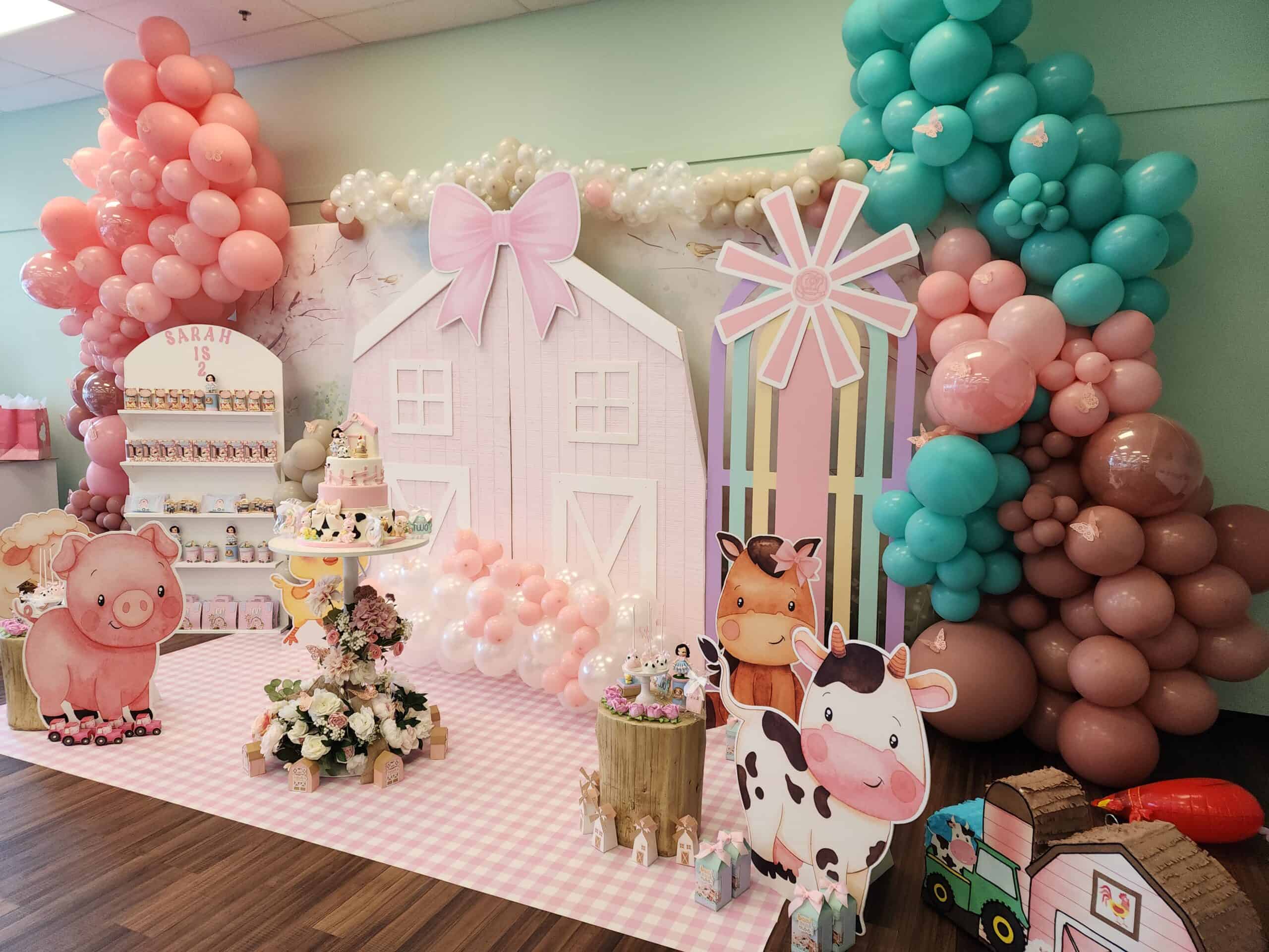 A farm-themed birthday party setup with pastel decorations, including a large pink barn backdrop adorned with a bow, colorful balloon garlands in shades of pink, teal, and brown, and cardboard cutouts of a pig, cow, and horse. The centerpiece features a tiered cake with farm animal decorations, surrounded by floral arrangements, party favors, and a sign that reads 'Sarah is 2' displayed on a candy shelf.